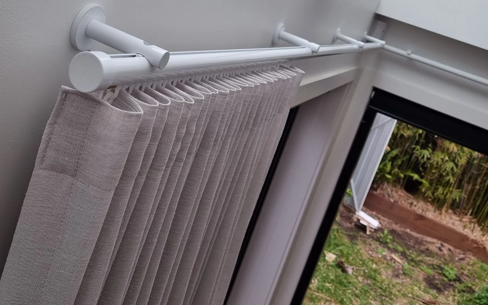 Close-up of a beige pleated curtain hanging on a white curtain rod above a window, with a view of a garden outside.