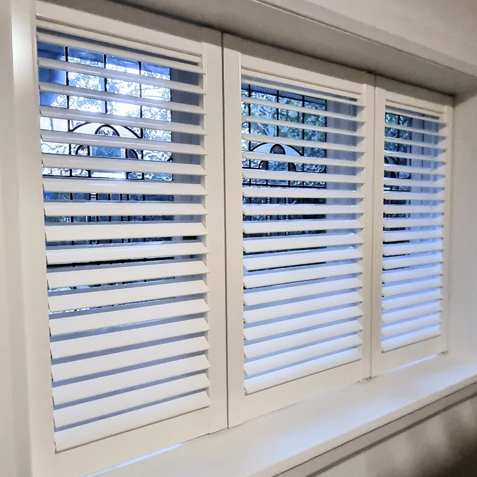 White window shutters with horizontal slats, partially opened, revealing trees and blue sky outside.