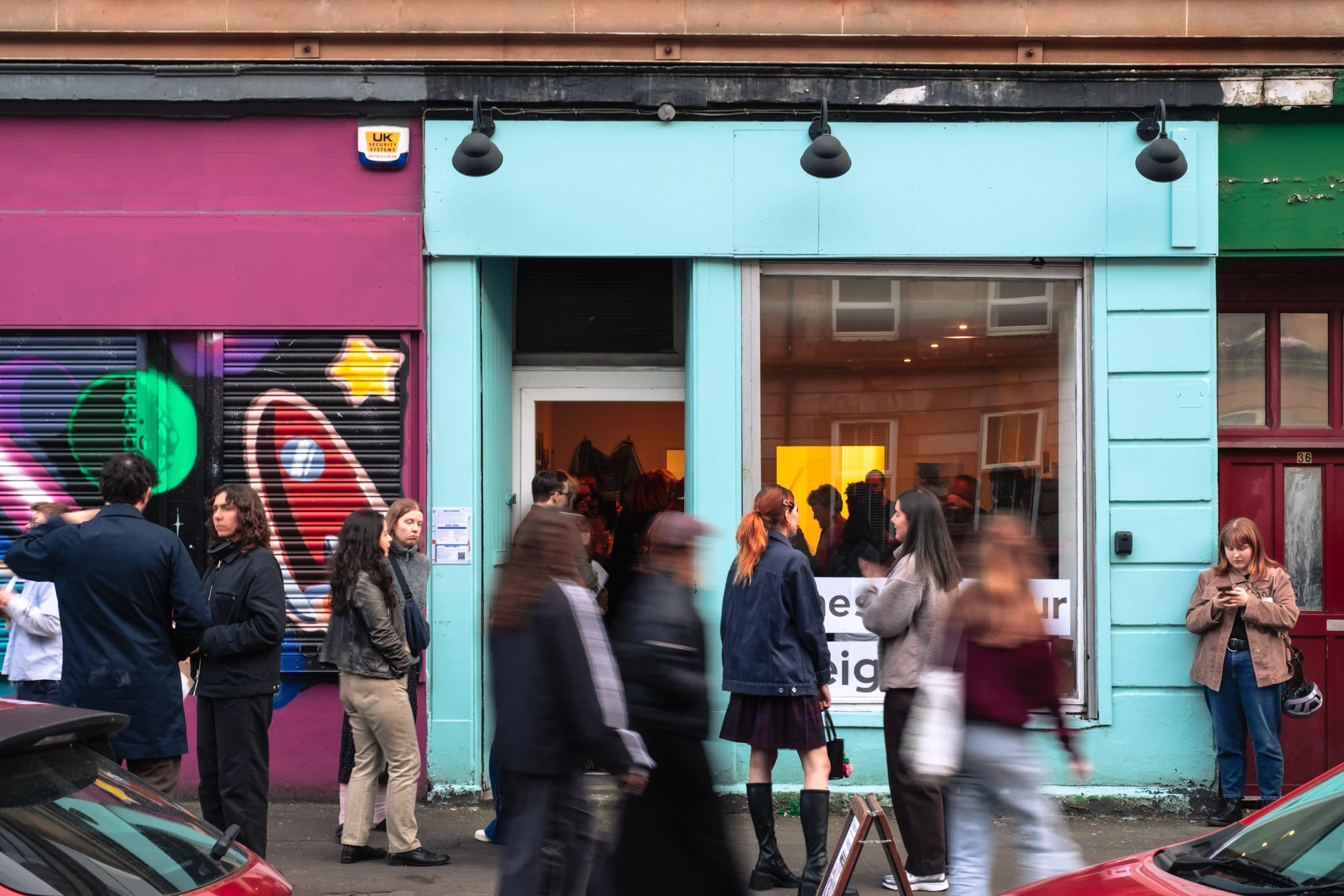 A busy street scene with people walking past a storefront featuring a turquoise and pink facade. Some individuals are blurred due to motion, while others stand still, some using their phones.