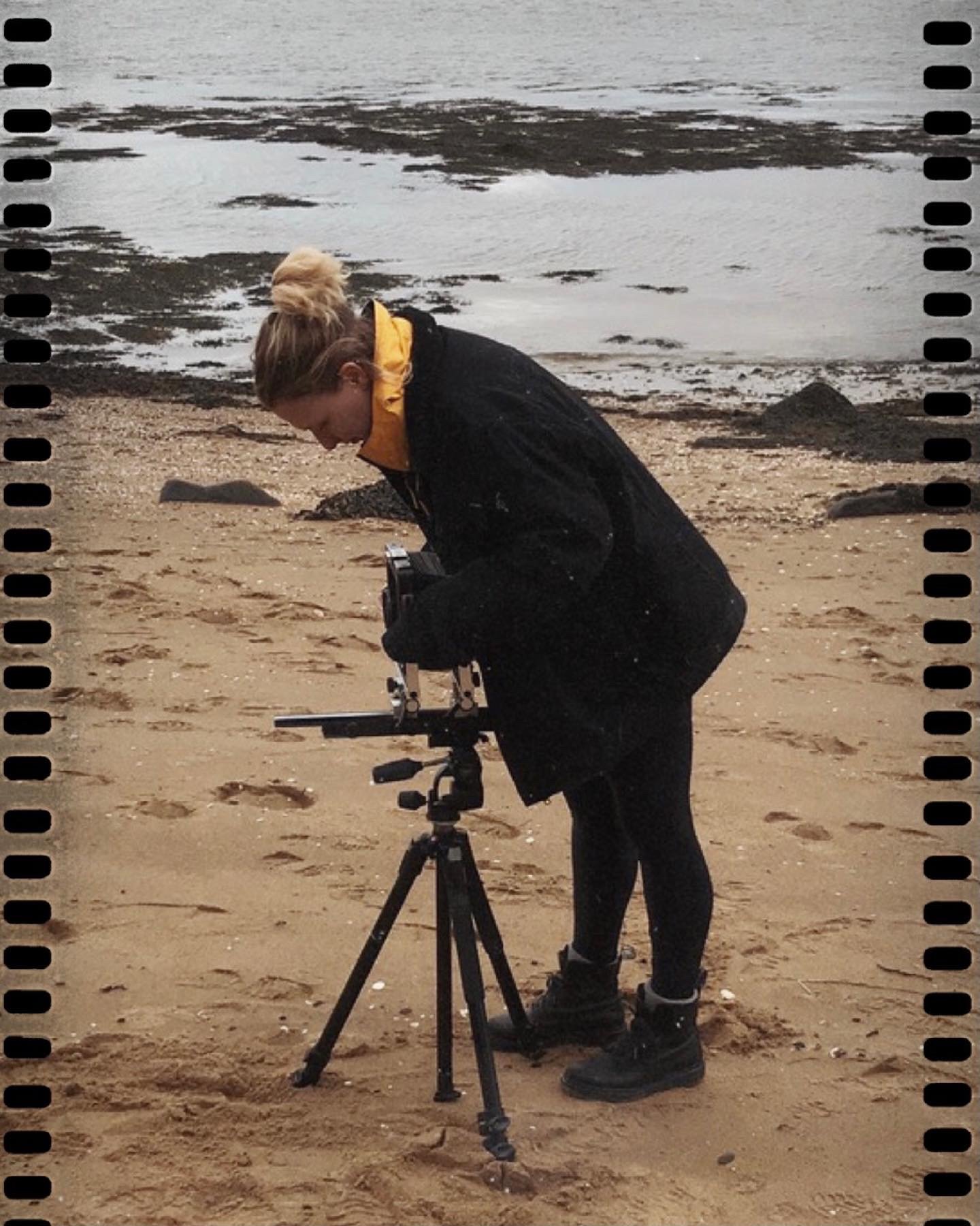 A person in outdoor clothing operating a camera on a tripod at a beach with sand, rocks, and water in the background.