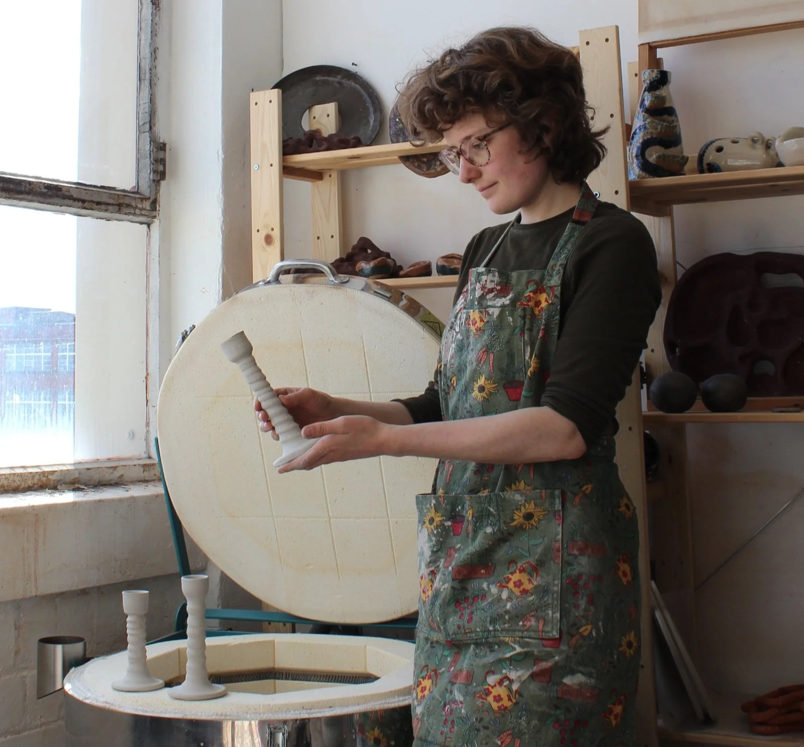 A person with curly hair and glasses wearing a floral apron, holding a pottery tool, standing in a pottery studio near a pottery wheel with some ceramic pieces and shelves with pottery in the background.
