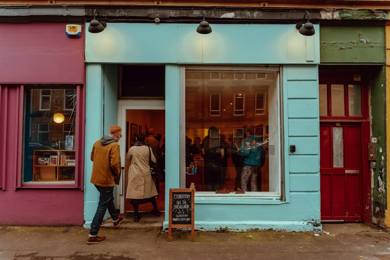 People entering a colorful art gallery with a blackboard sign outside, painted in pink, blue, and green, on a city sidewalk.