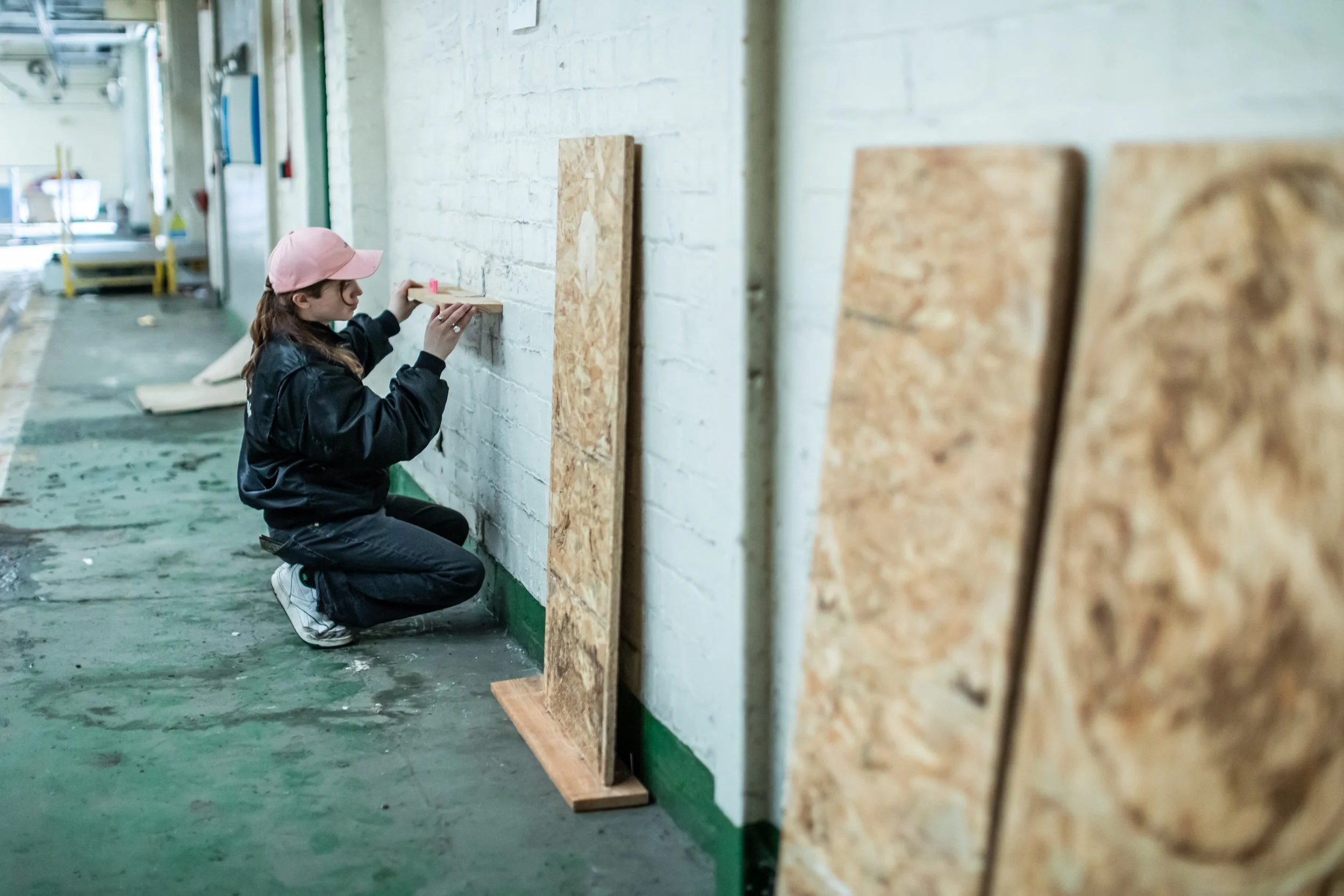 A young woman in a pink cap and black jacket kneels on the floor and marks a piece of wood with a pink marker in a workshop with a white brick wall and plywood boards leaning against the wall.