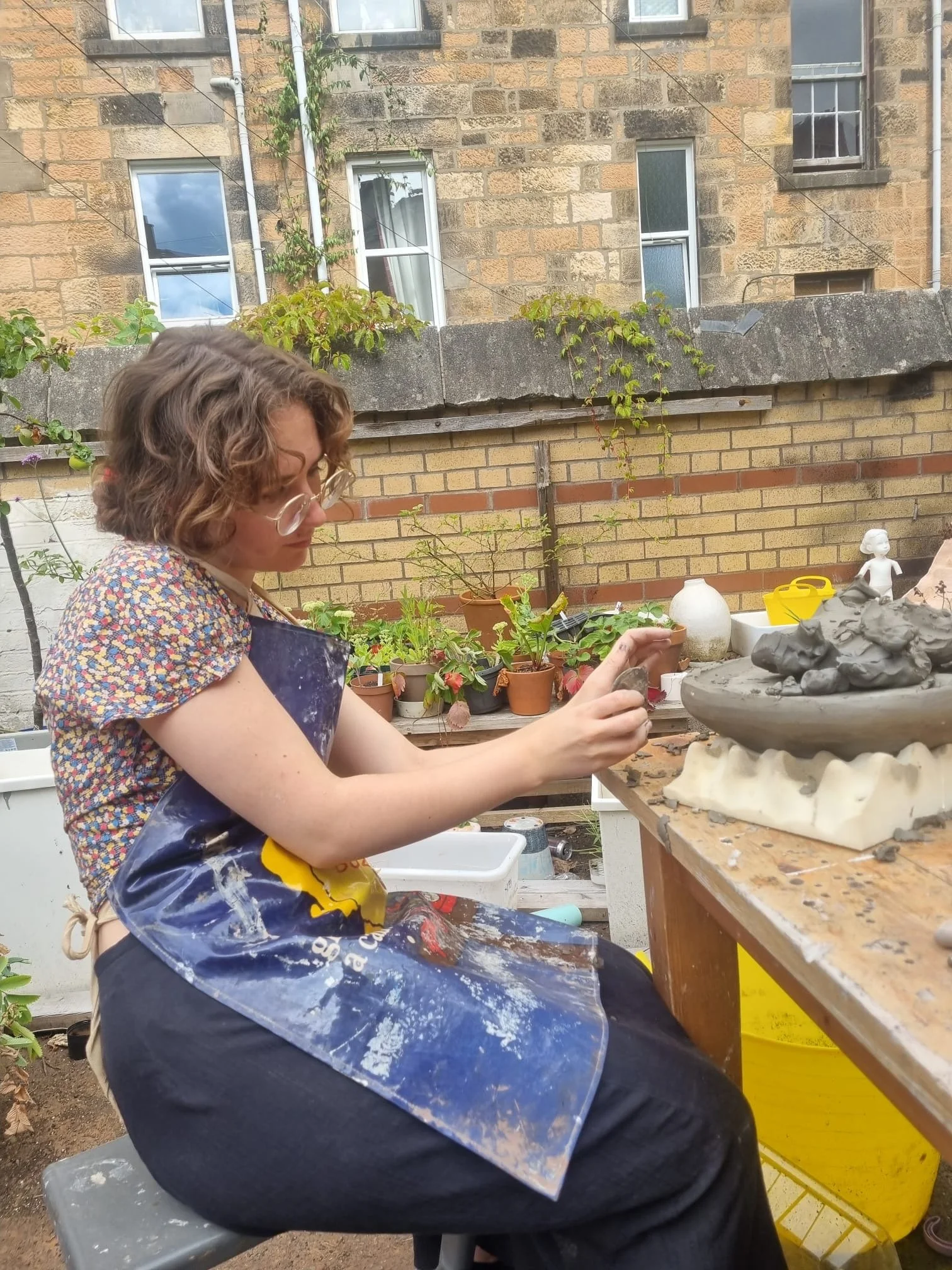 A woman working on a pottery sculpture outdoors, surrounded by potted plants and gardening tools, with a brick wall and windows in the background.