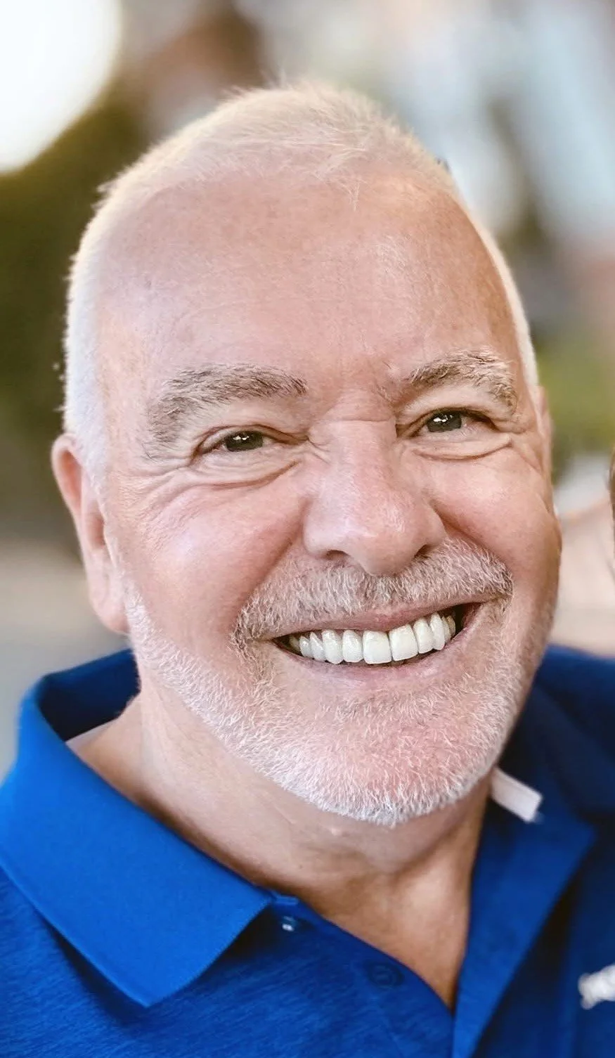 Close-up of a smiling older man with white hair, a beard, and wearing a blue shirt.