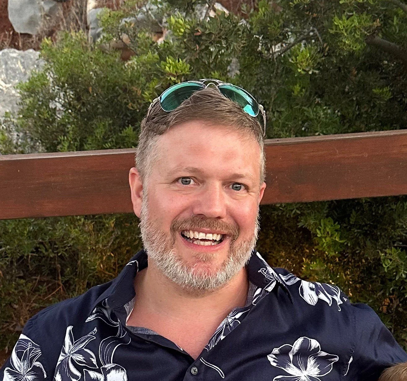 A smiling man with gray hair and a beard wearing sunglasses on his head and a navy blue Hawaiian shirt with white floral patterns, outdoors with greenery and a wooden rail in the background.