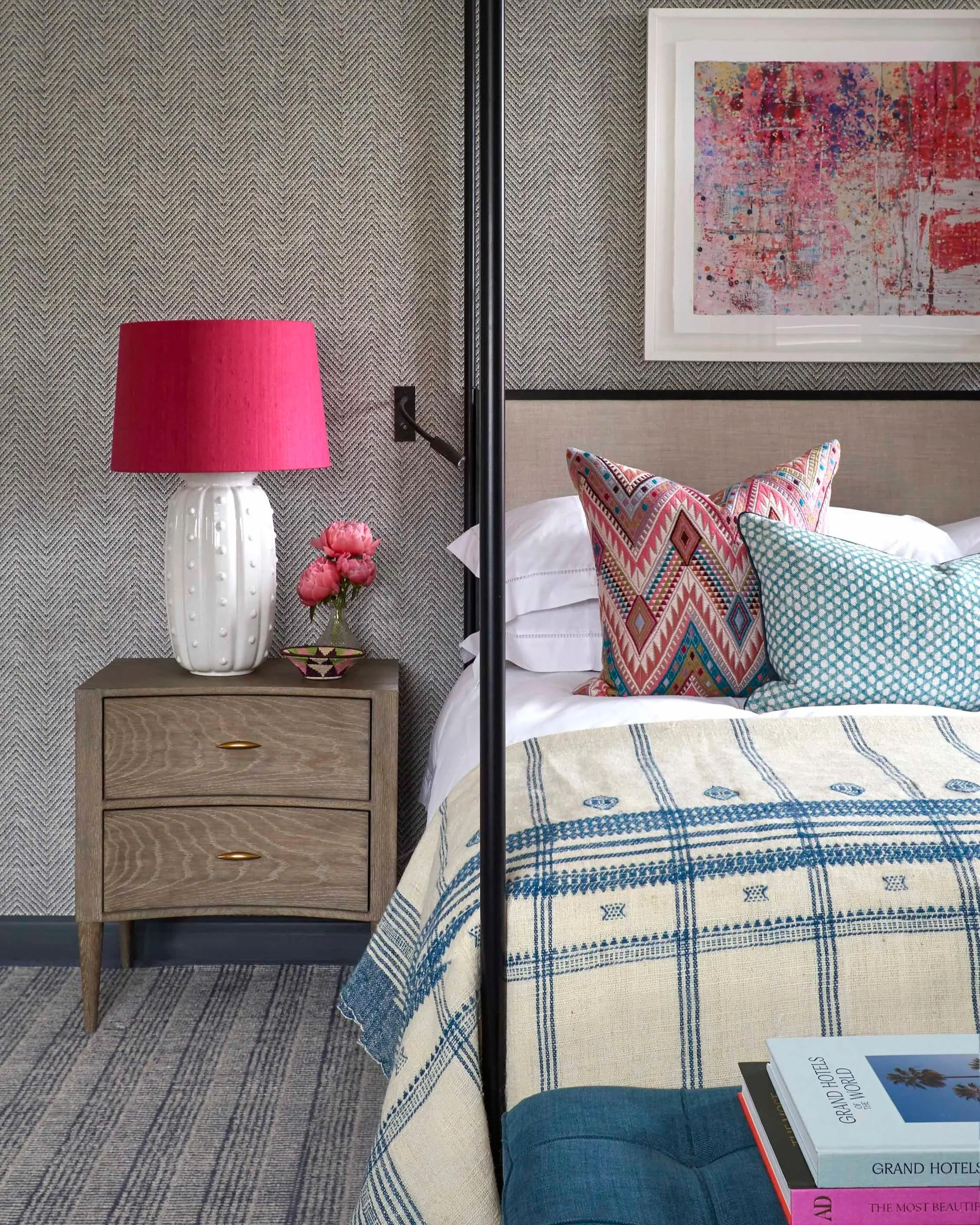 A hotel room with a beige headboard, decorative throw pillows, a bed with a blue and white patterned duvet, a wooden nightstand with a white vase holding pink flowers, and a pink lampshade on a white table lamp. Framed abstract artwork hangs on the w