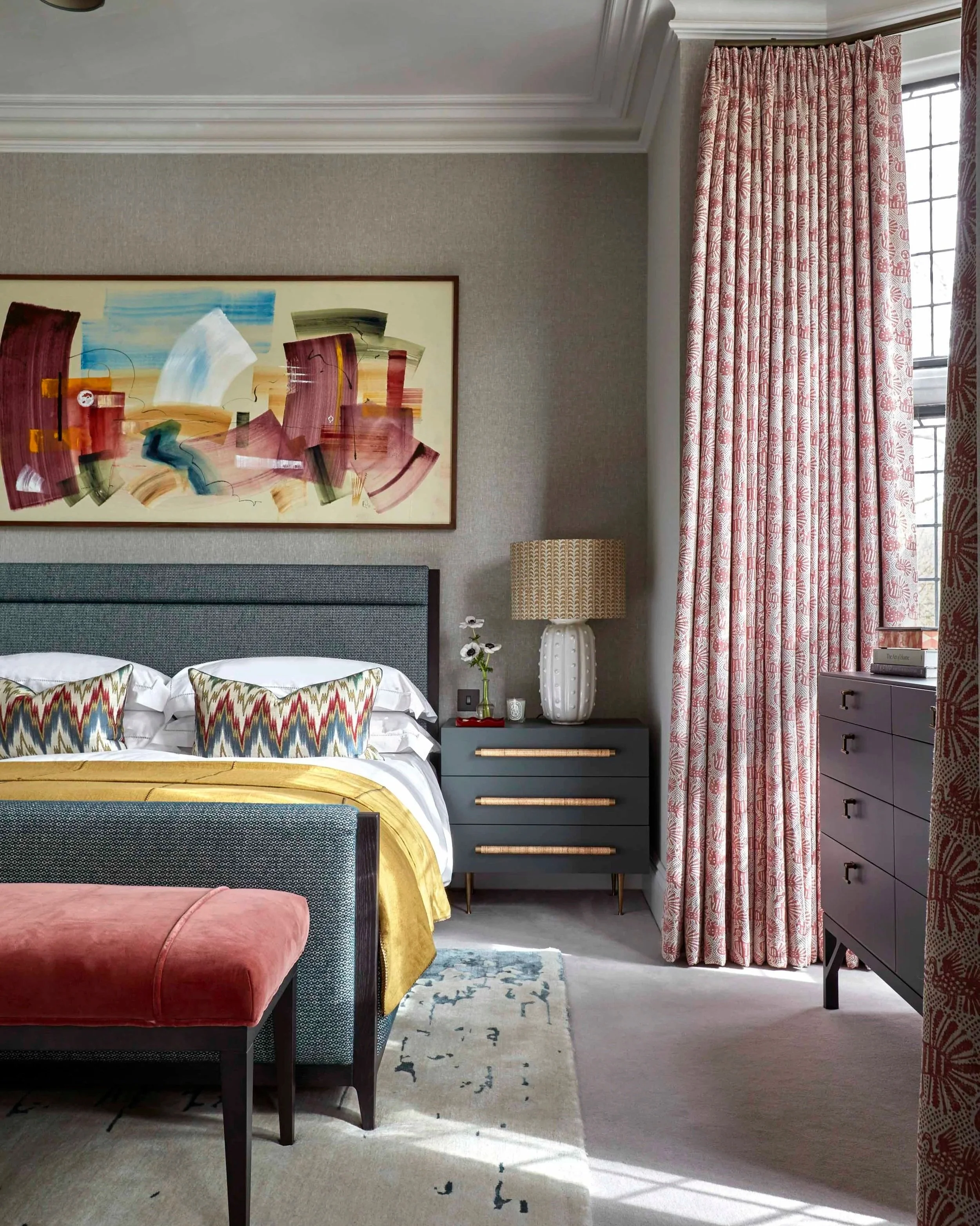 A bedroom with a bed, a dark gray nightstand with gold handles, a white vase with black and white flowers, a lamp, and abstract artwork on the wall. There are pink and white patterned curtains next to a window with black framing. The room has a beige carpet and a light-colored wall.