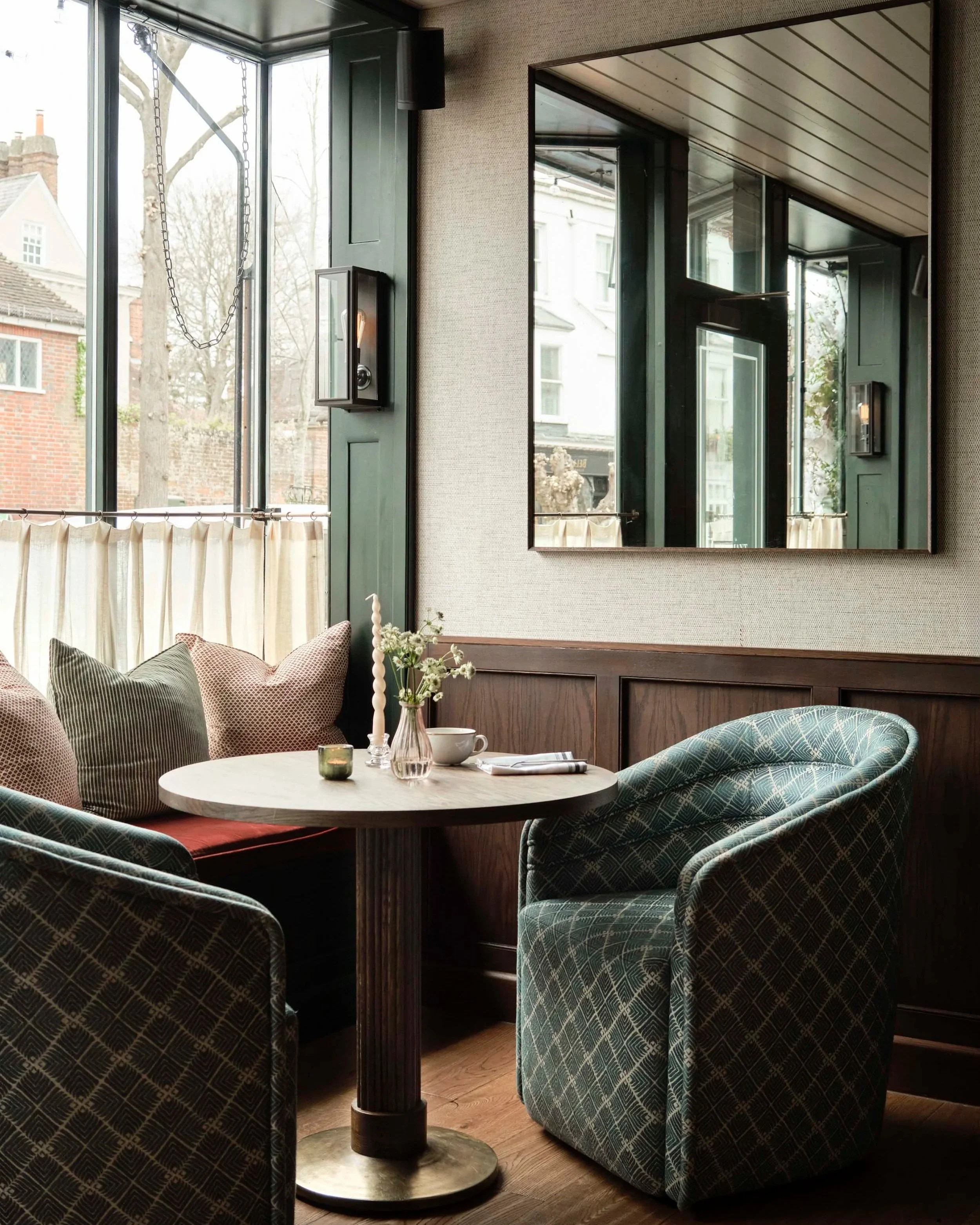Le Flamant Restaurant and Wine Bar, Sussex. Interior of a cozy cafe or restaurant with a round table and upholstered chairs. A small vase with flowers and a candle are on the table. Large windows let in natural light, showing trees and buildings outs