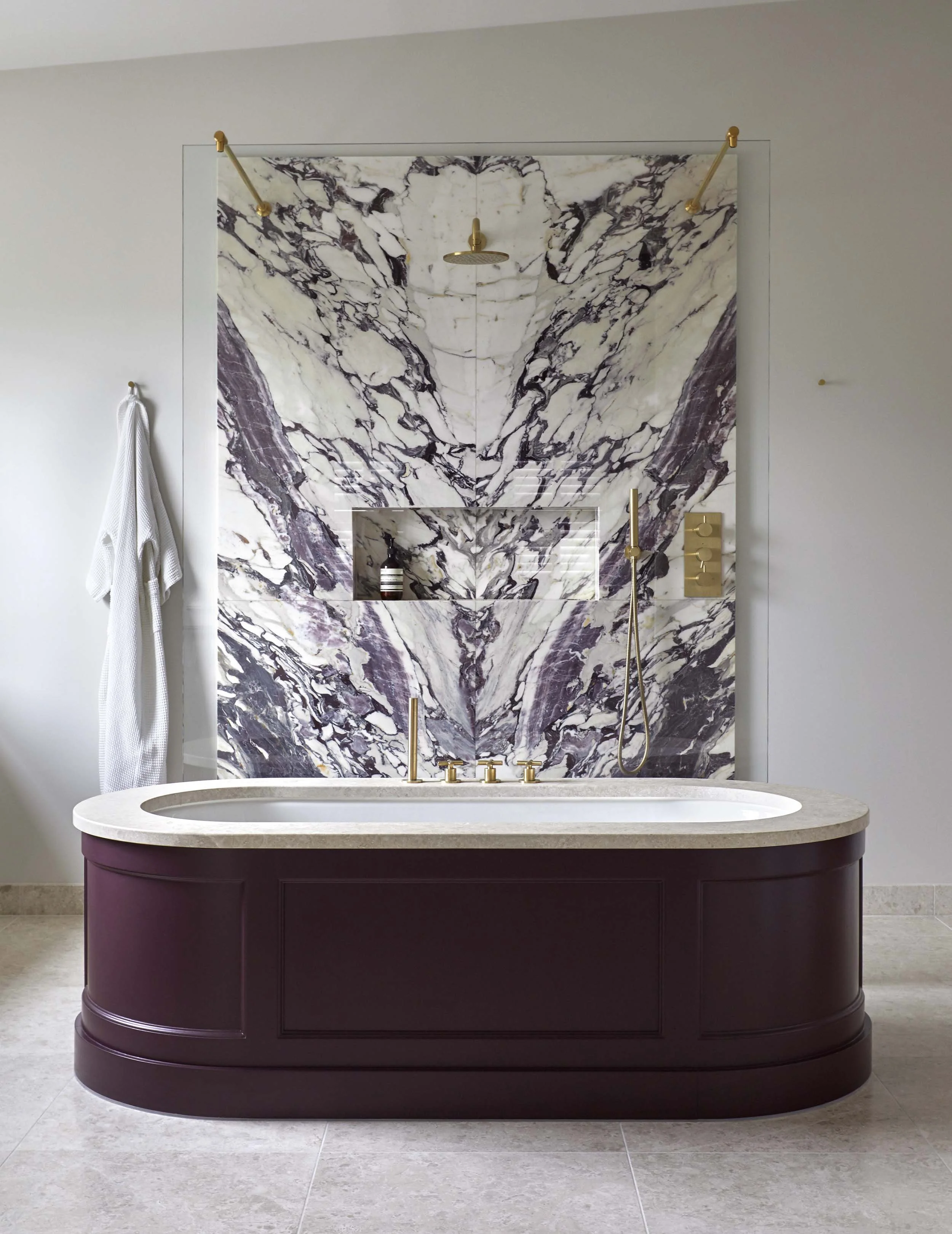 Bathroom with a purple vanity, marble wall, and a bathtub with a beige countertop. The marble wall has a showerhead and a niche with toiletries. There is a white towel hanging on a hook on the wall.