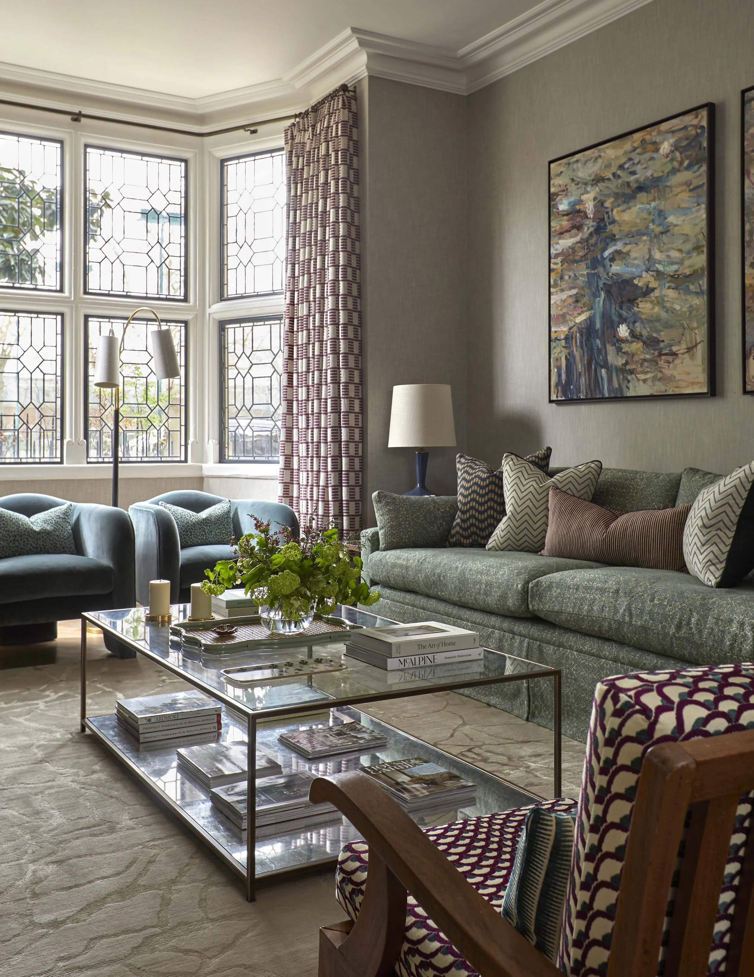 Living room with large bay window, patterned curtains, green sofa with decorative pillows, glass coffee table with books and a floral arrangement, beige area rug, abstract painting on wall, floor lamp, and colorful armchair.