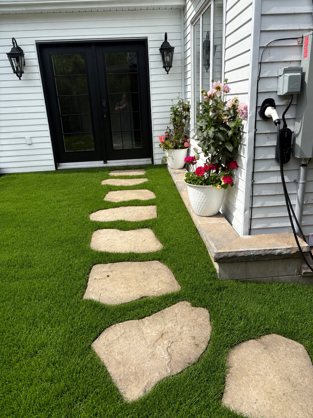 Chemin de pierre menant à une porte d'entrée noire, entouré d'herbe verte et de pots de fleurs colorées, avec un mur de maison en revêtement blanc.