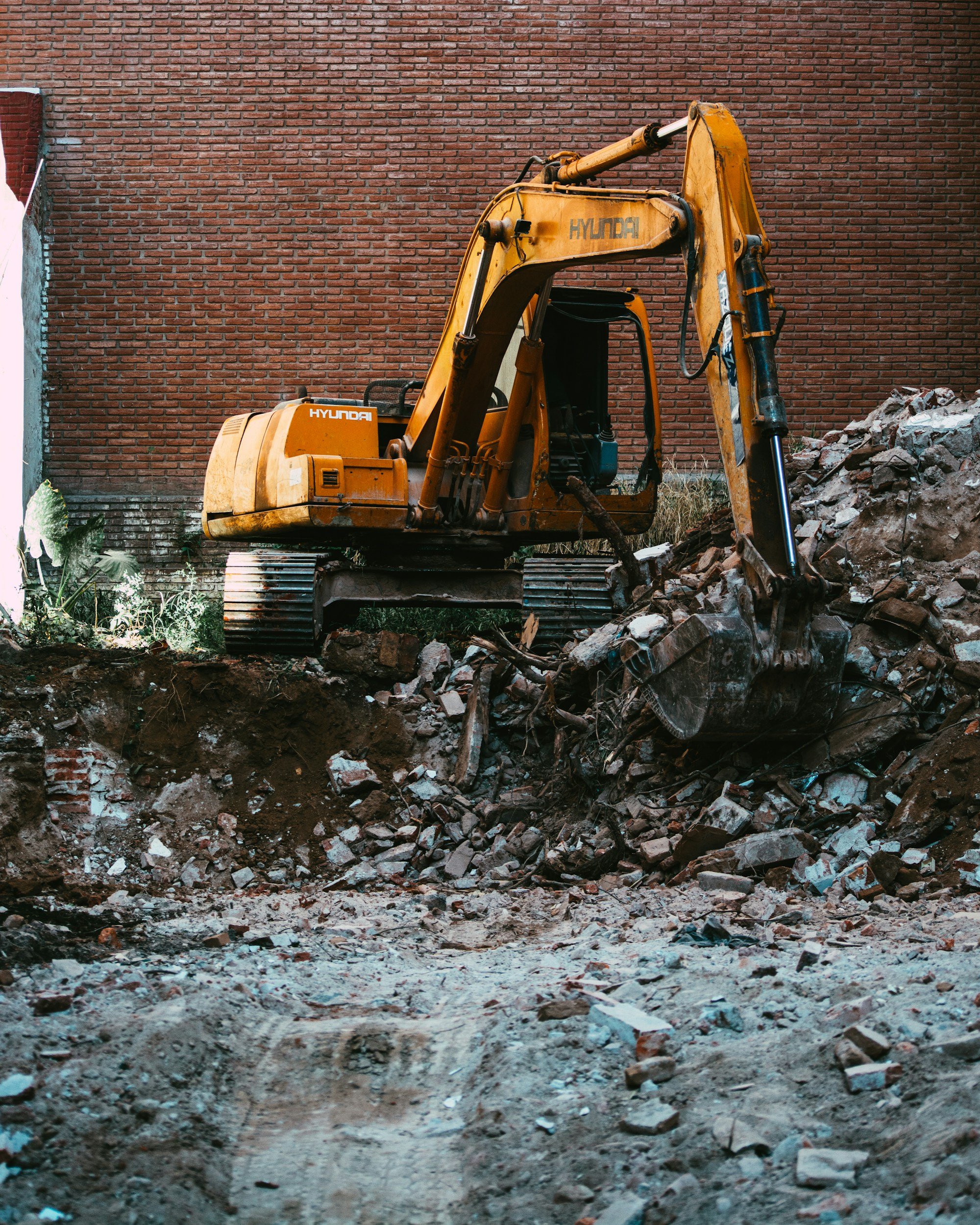 Une pelleteuse jaune en construction à côté d’un mur de briques rouges, entourée de débris de construction et de terre.