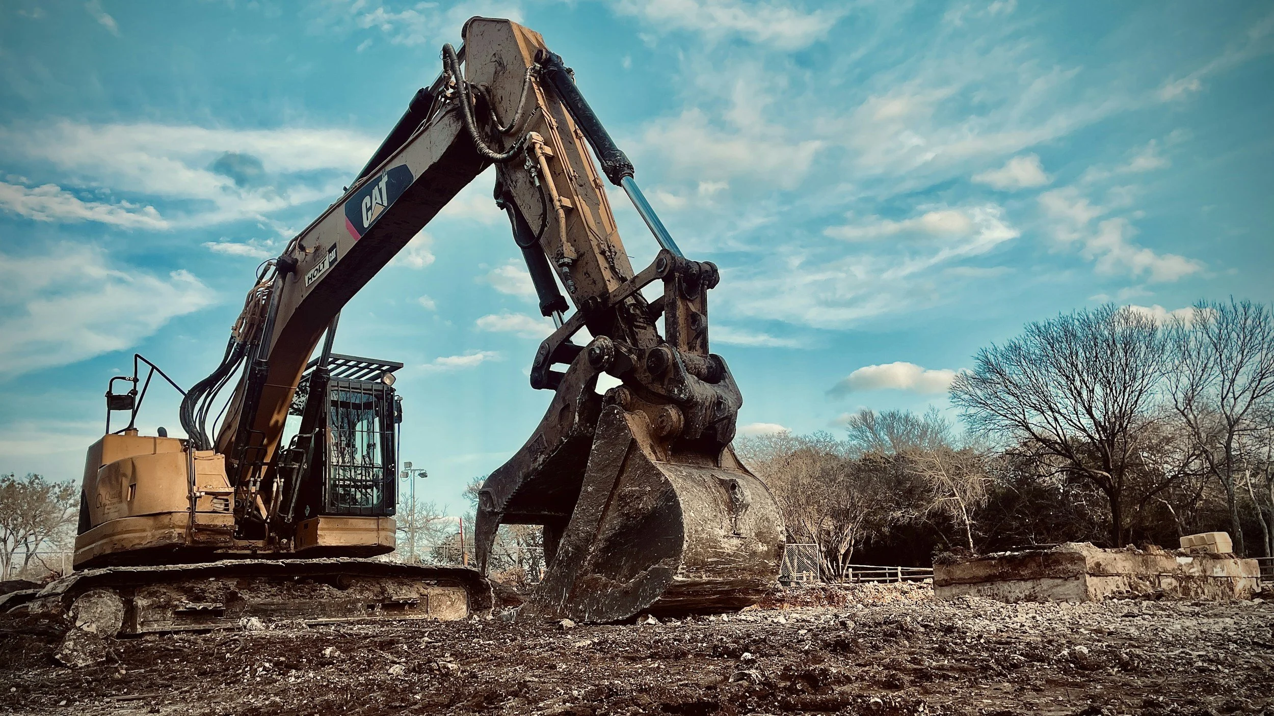 Une pelleteuse en activité sur un site de construction avec un ciel bleu et quelques nuages, et des arbres en arrière-plan.
