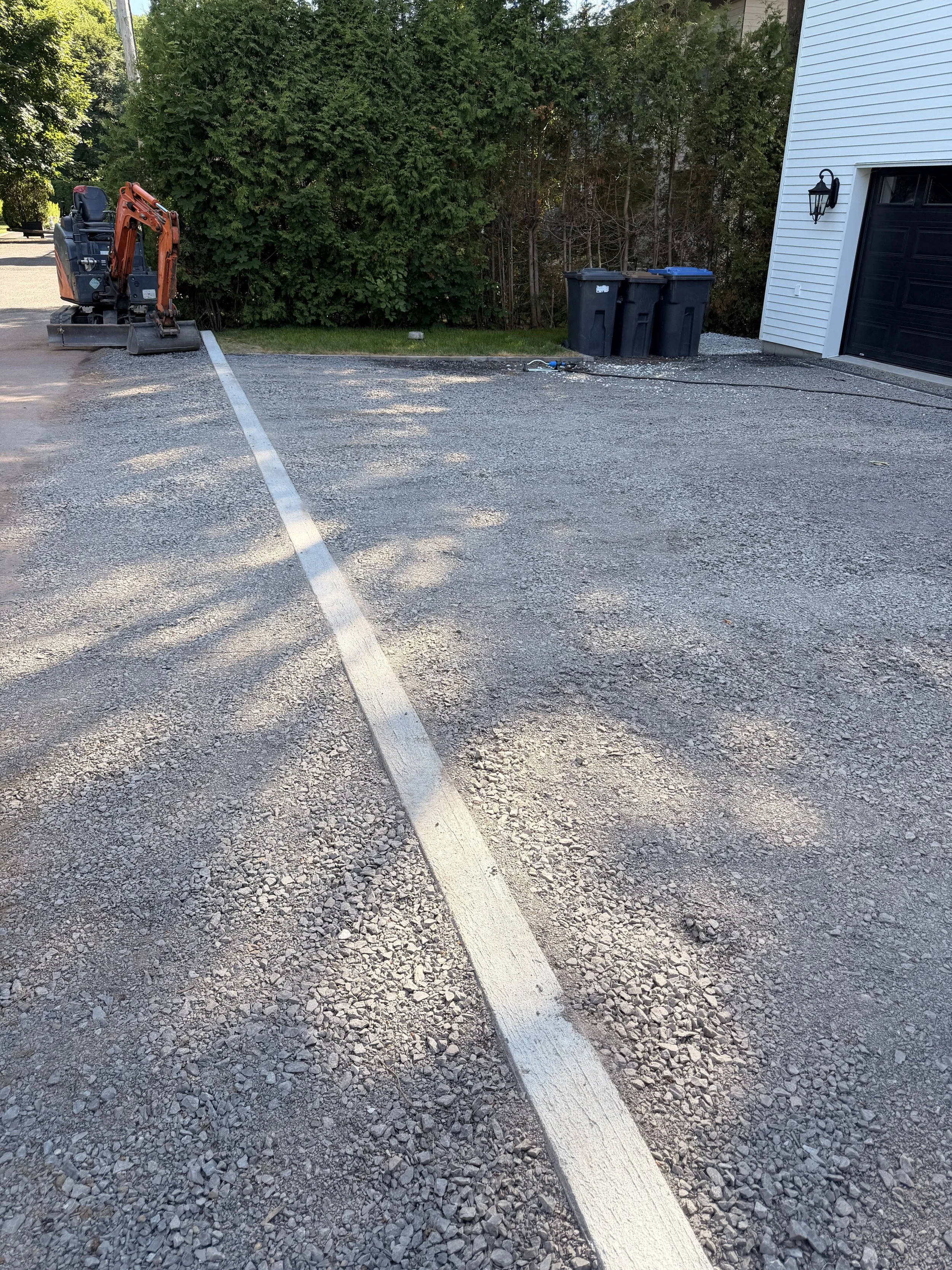 Un espace de stationnement en construction avec un mini-pelle orange, trois bacs poubelle noirs, un câble électrique et une maison blanche avec une porte de garage noire.