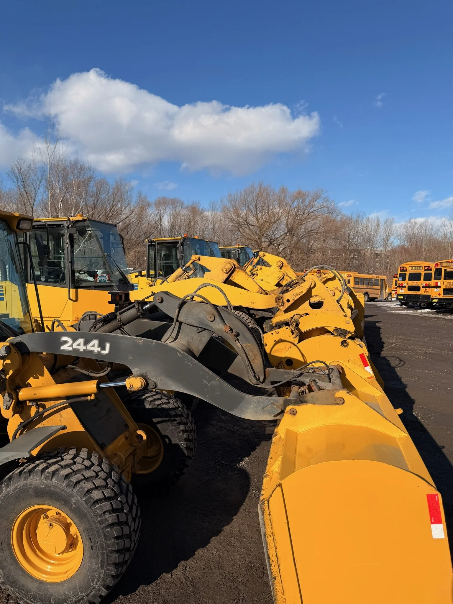 Ligne de bulldozers jaunes alignés dans un parking extérieur avec des bus scolaires rouges à l'arrière-plan, ciel bleu avec quelques nuages, arbres sans feuilles.