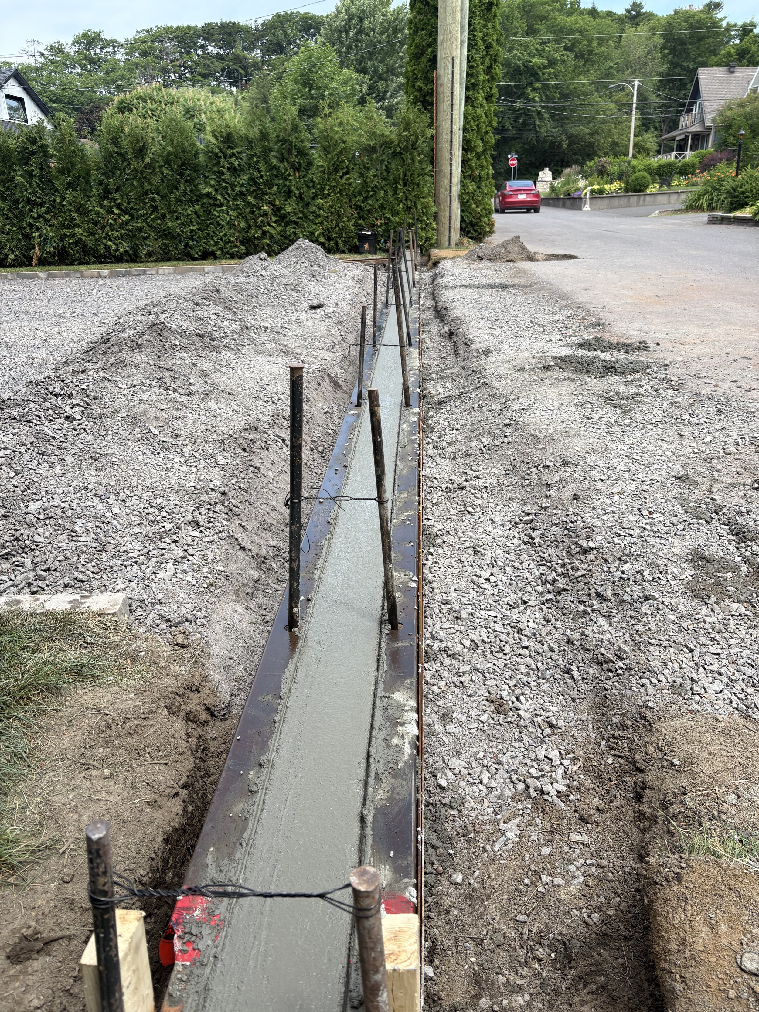 Une allée en construction pavée en béton, entourée de gravier et de terre, avec des poteaux en bois et un cadre métallique pour soutenir la bétonnière, dans une zone résidentielle avec des maisons et des arbres en arrière-plan.