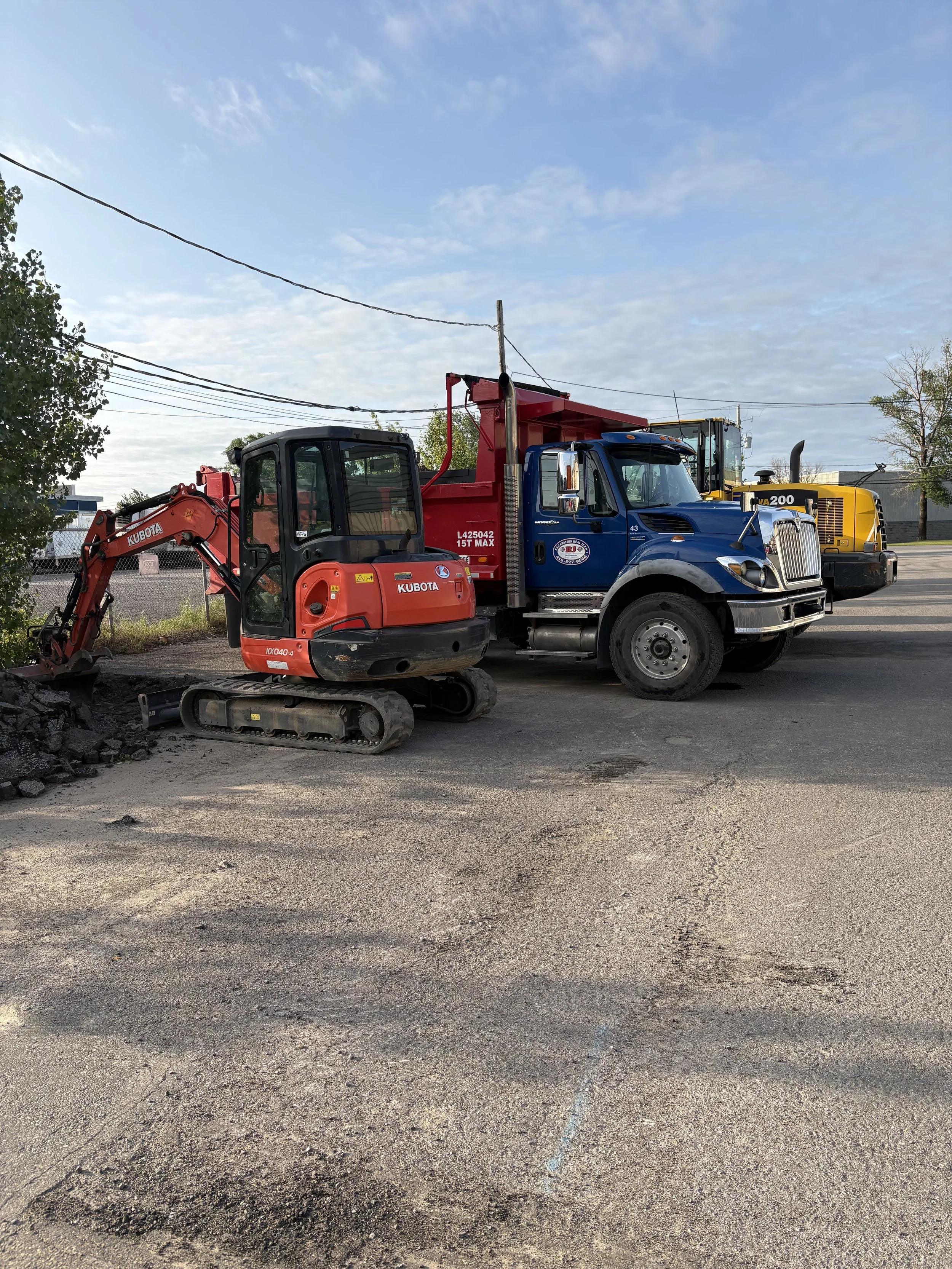 Construction site with a small red excavator and a large blue and yellow road maintenance truck on a paved area under a partly cloudy sky.