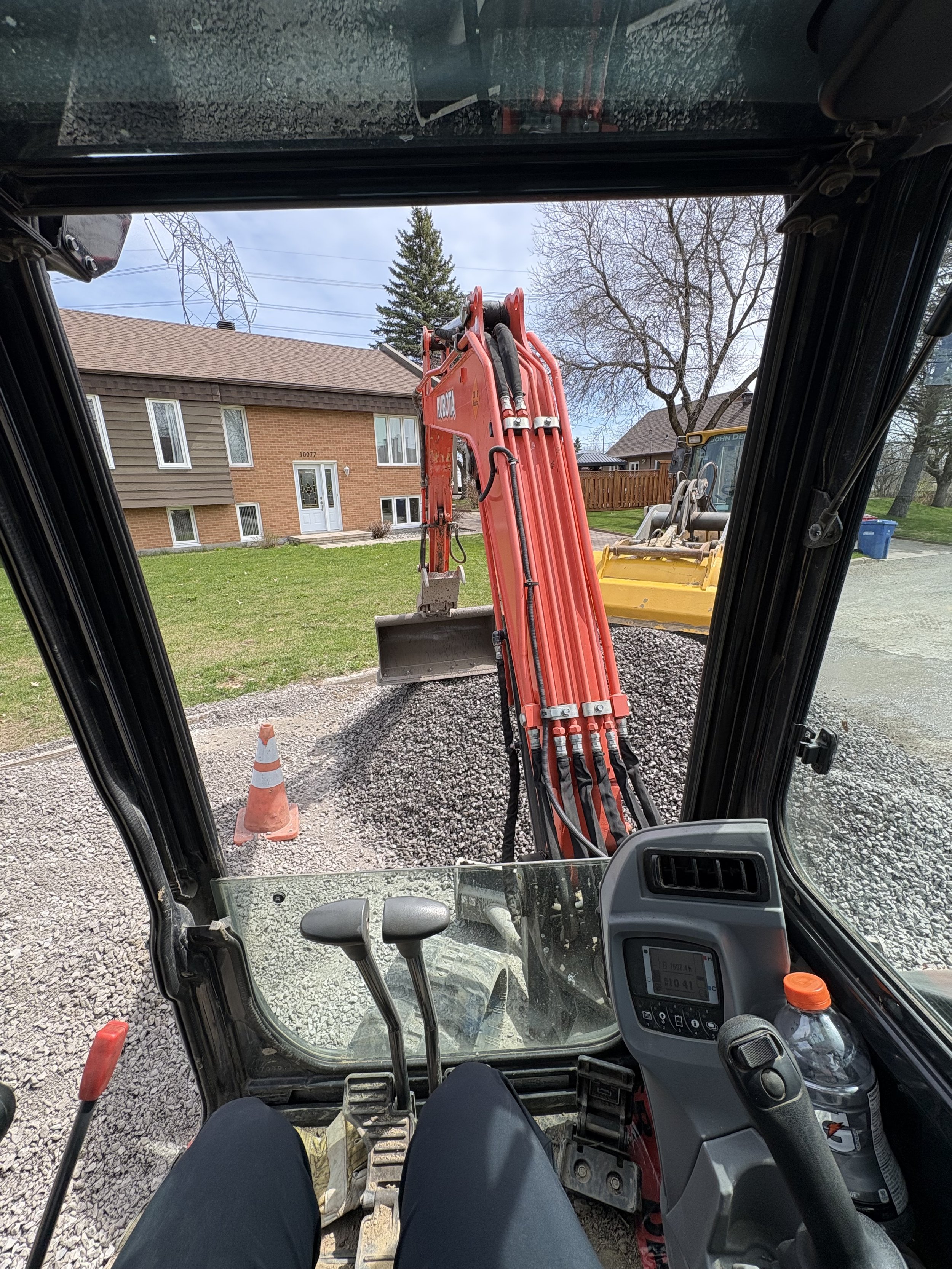 Vue depuis l'intérieur d'une excavatrice ou machine de construction, regardant vers l'extérieur, avec un bâtiment résidentiel, un arbre, une cabine de machine, un panneau de trafic et du matériel de construction visible dans le cadre.