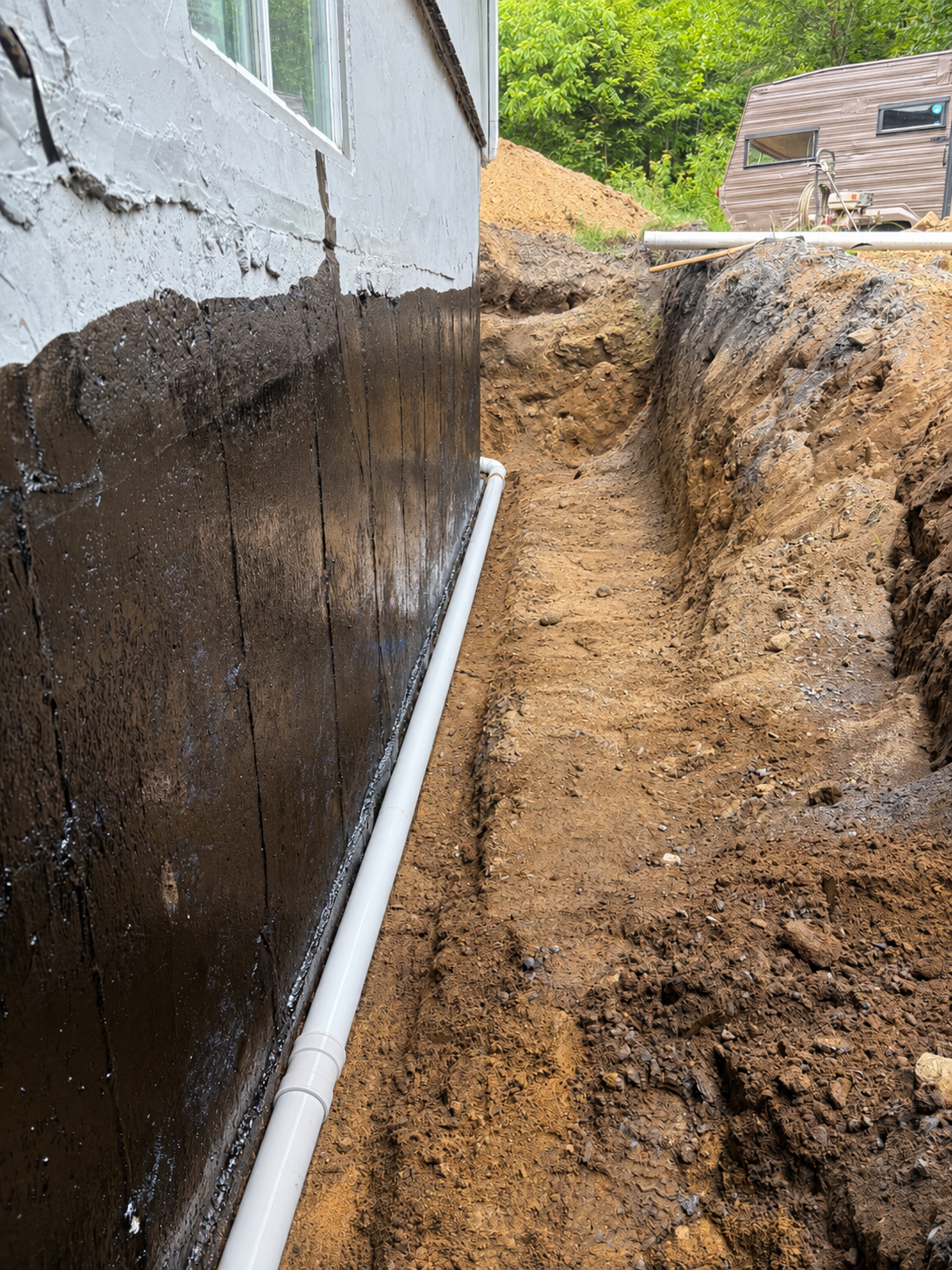 Installation de plomberie avec un tuyau en PVC le long du fond d'une tranchée creusée à côté d'une maison, avec un revêtement noir sur la maison et un caravane en arrière-plan.