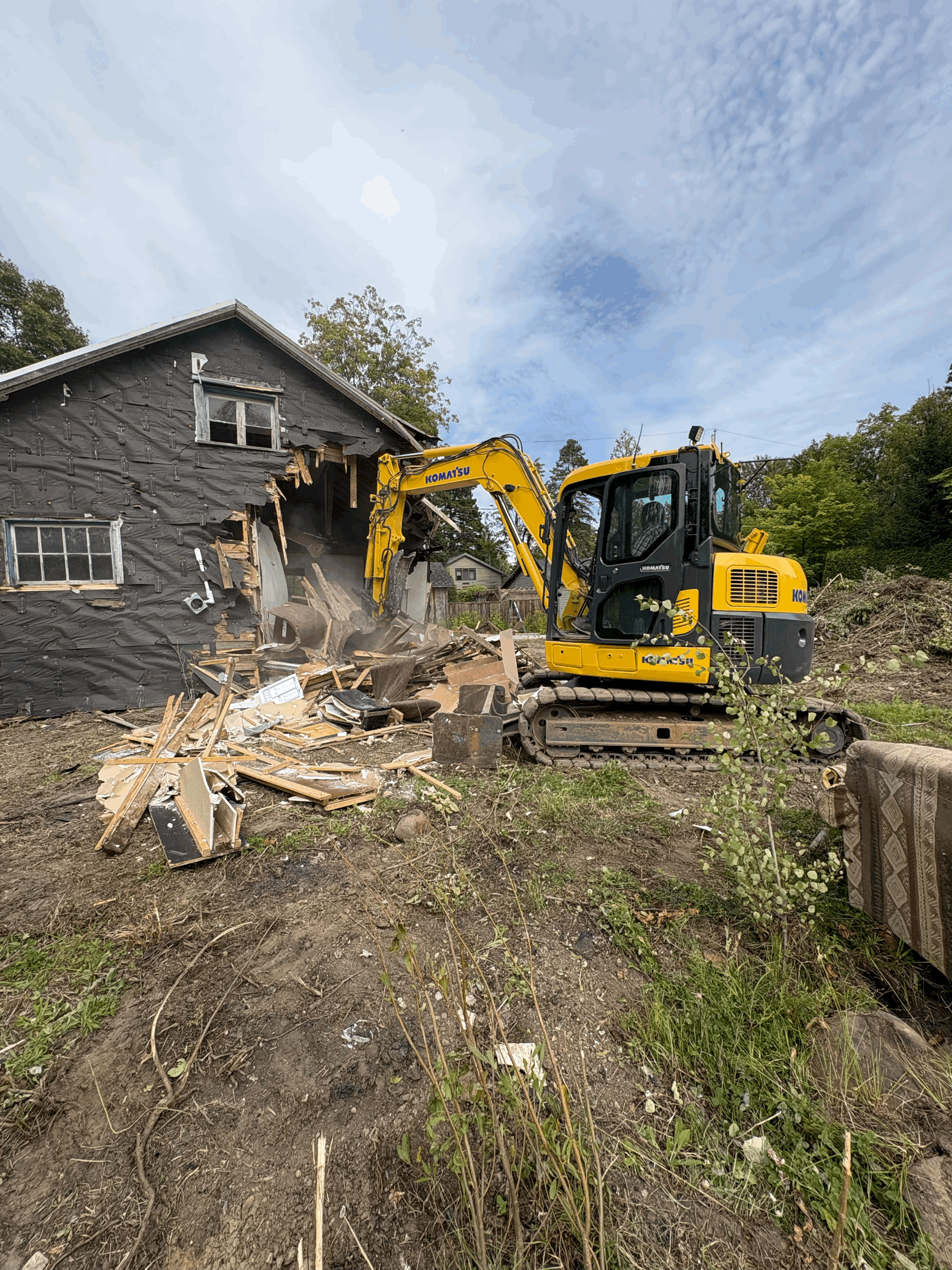 Une maison en cours de démolition avec une pelleteuse jaune en action, déplaçant des débris, sous un ciel partiellement nuageux.