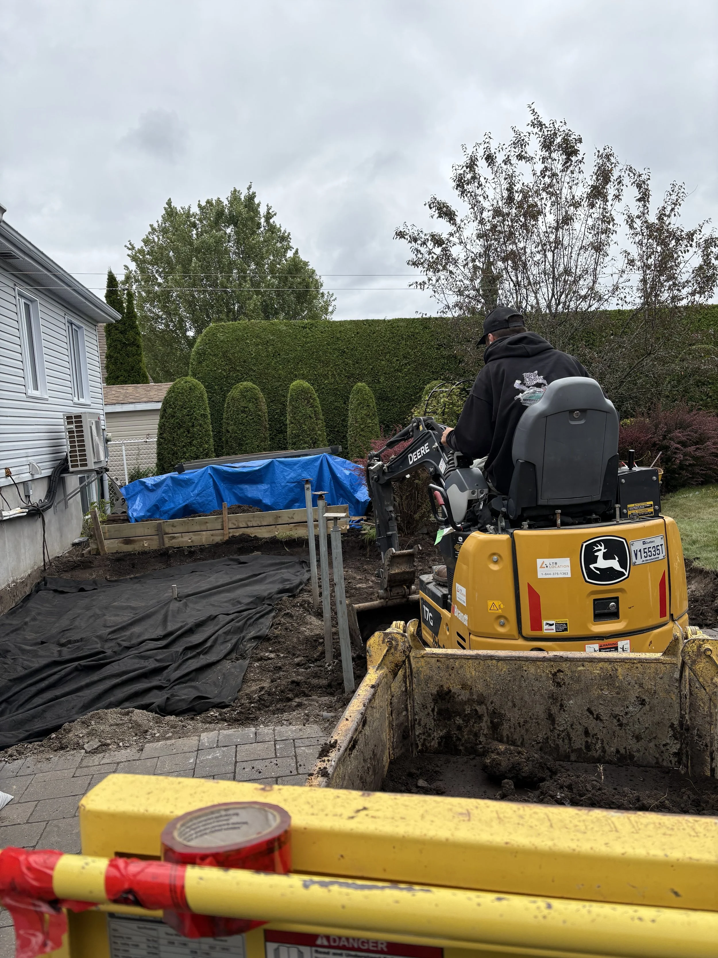 Une scène de chantier avec un homme travaillant sur une pelleteuse jaune JD, à l'arrière-plan un jardin avec des haies et des arbres, et un ciel nuageux.