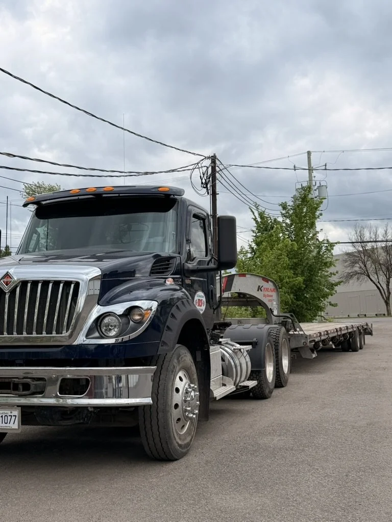 Un camion noir à plateforme allongée stationné sur une route, avec des lignes électriques visibles au-dessus et des arbres en arrière-plan sous un ciel nuageux.