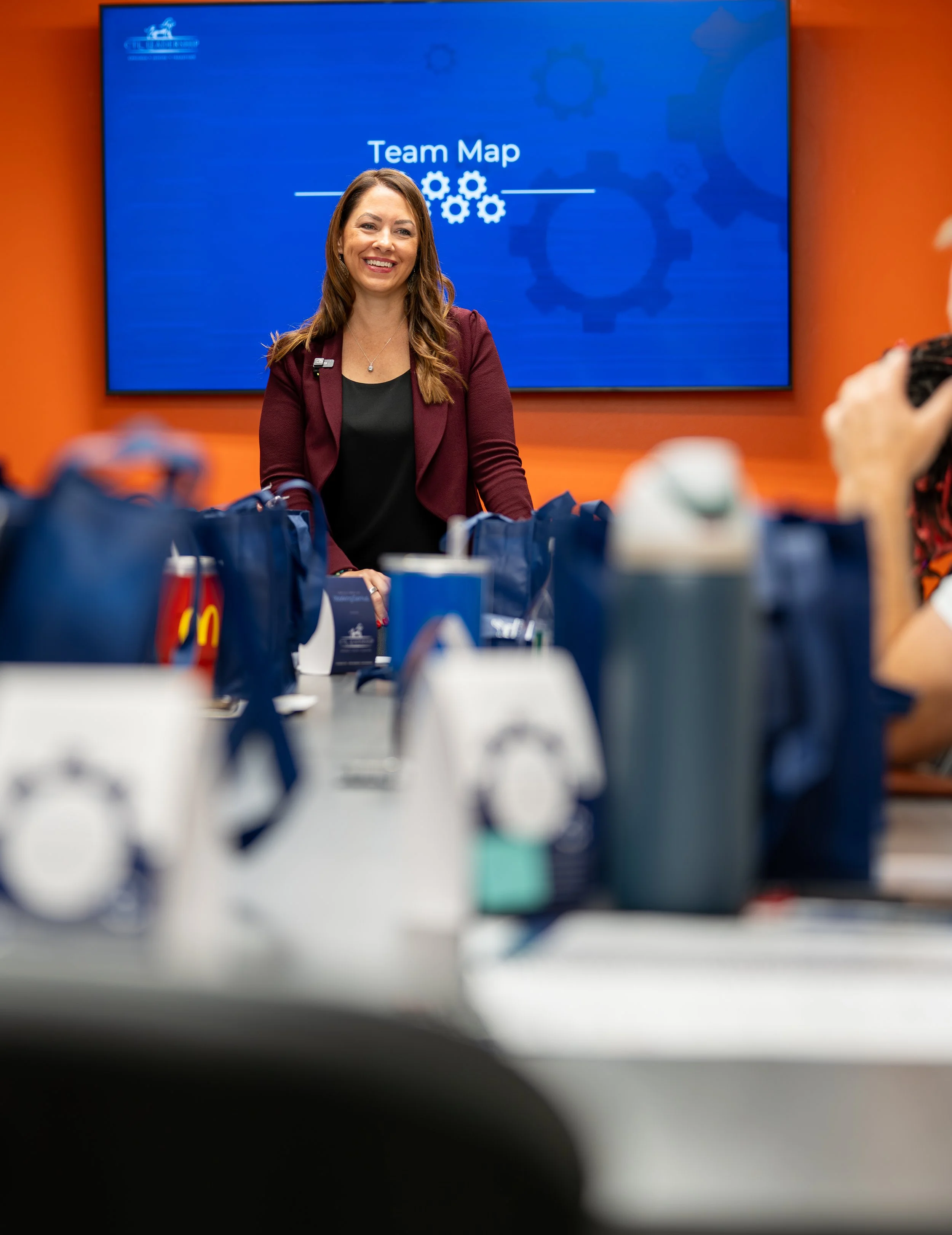 A woman with long brown hair smiling at a camera, sitting in front of a blue screen that reads 'Team Map' and has gear icons. The environment appears to be a conference or presentation room with a table and pitchers in the foreground.