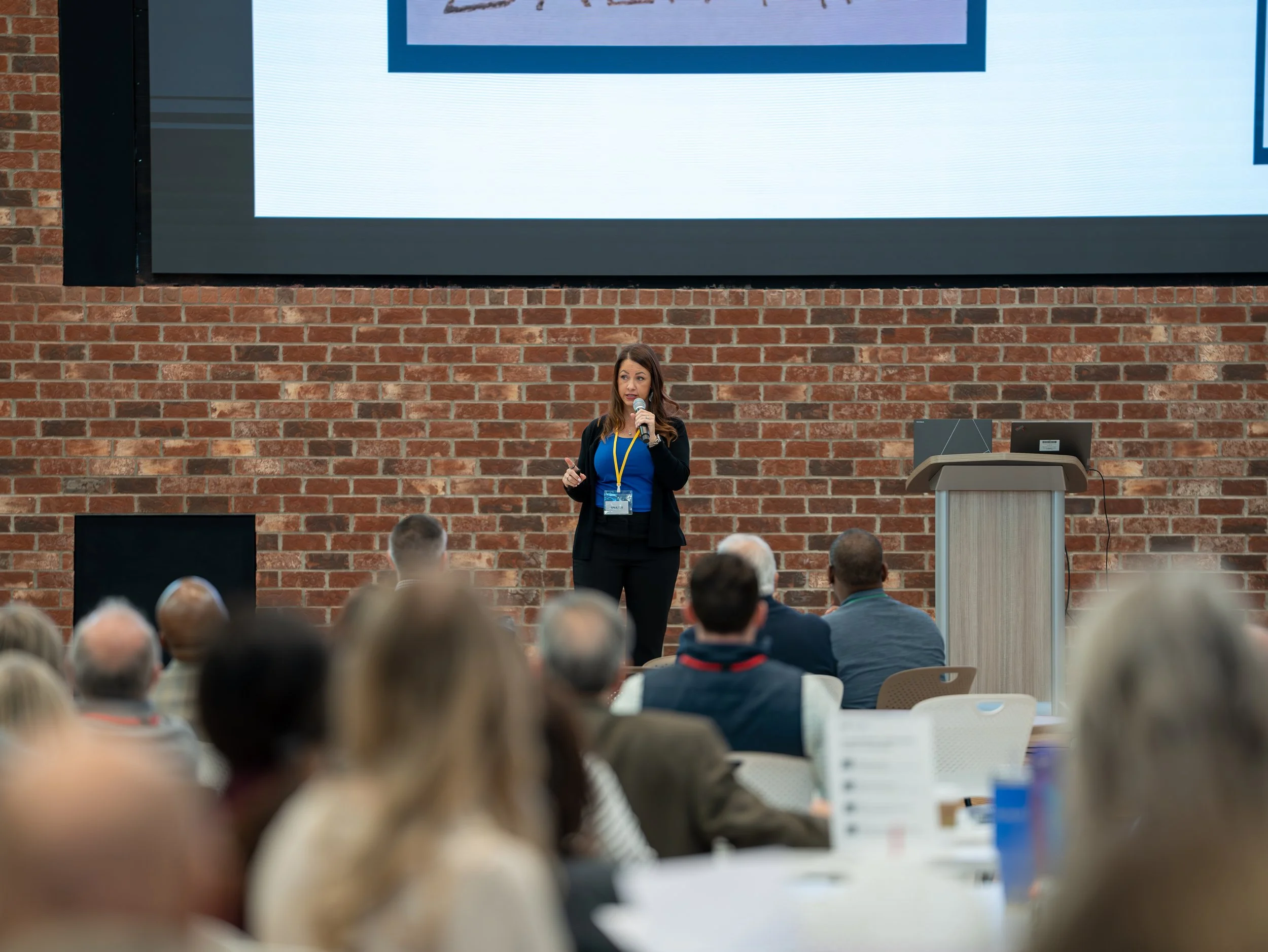 A woman with long brown hair, wearing a blue shirt and black blazer, is giving a presentation at a conference in front of a large audience. She is holding a microphone and a clicker. The presentation is displayed on a large screen behind her, mounted on a brick wall.