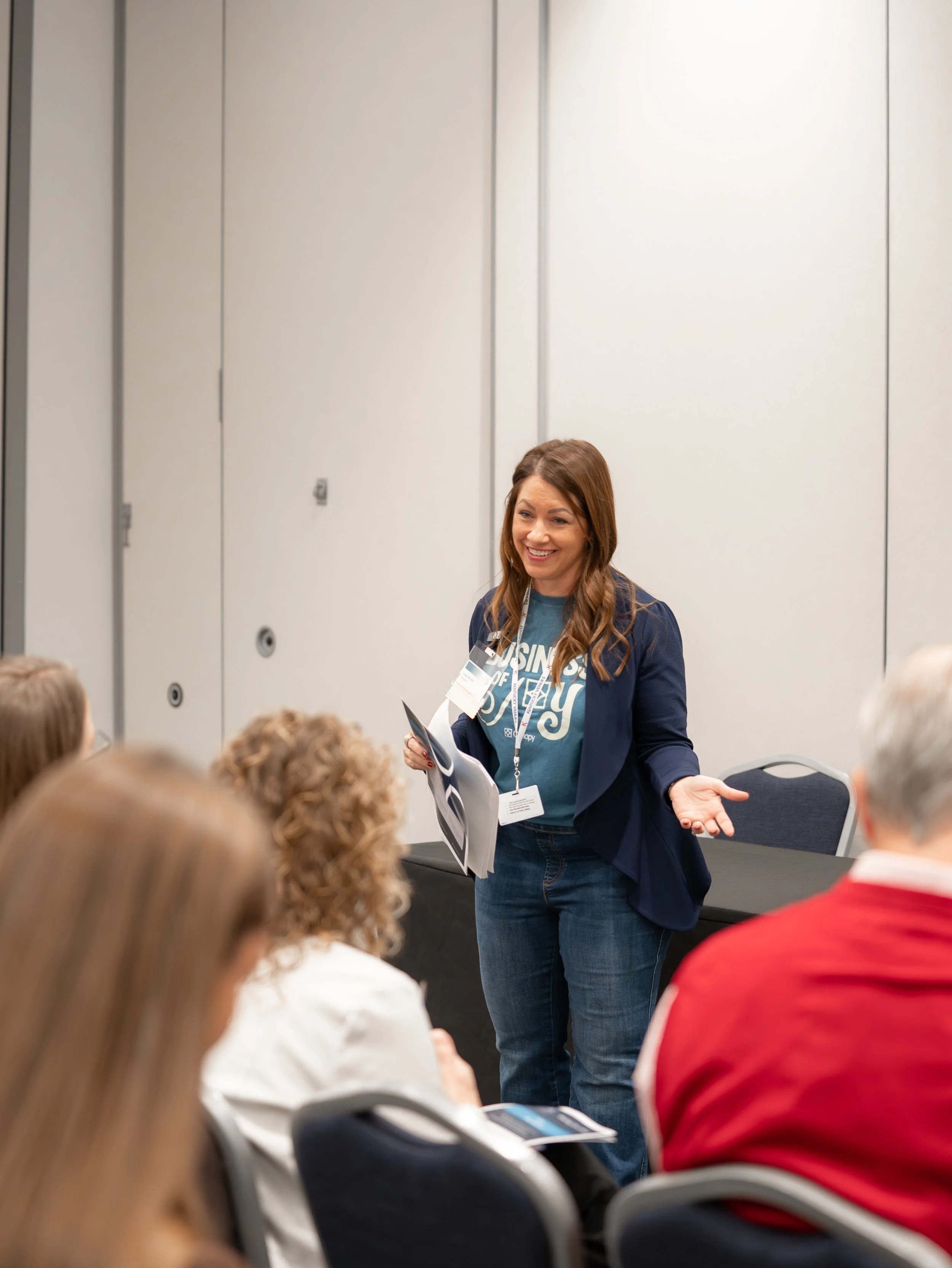 A woman giving a presentation in a conference room with an audience. She is holding papers and wearing a blue jacket and a blue T-shirt with text. The audience members are seated and facing her, some with headsets or earbuds.
