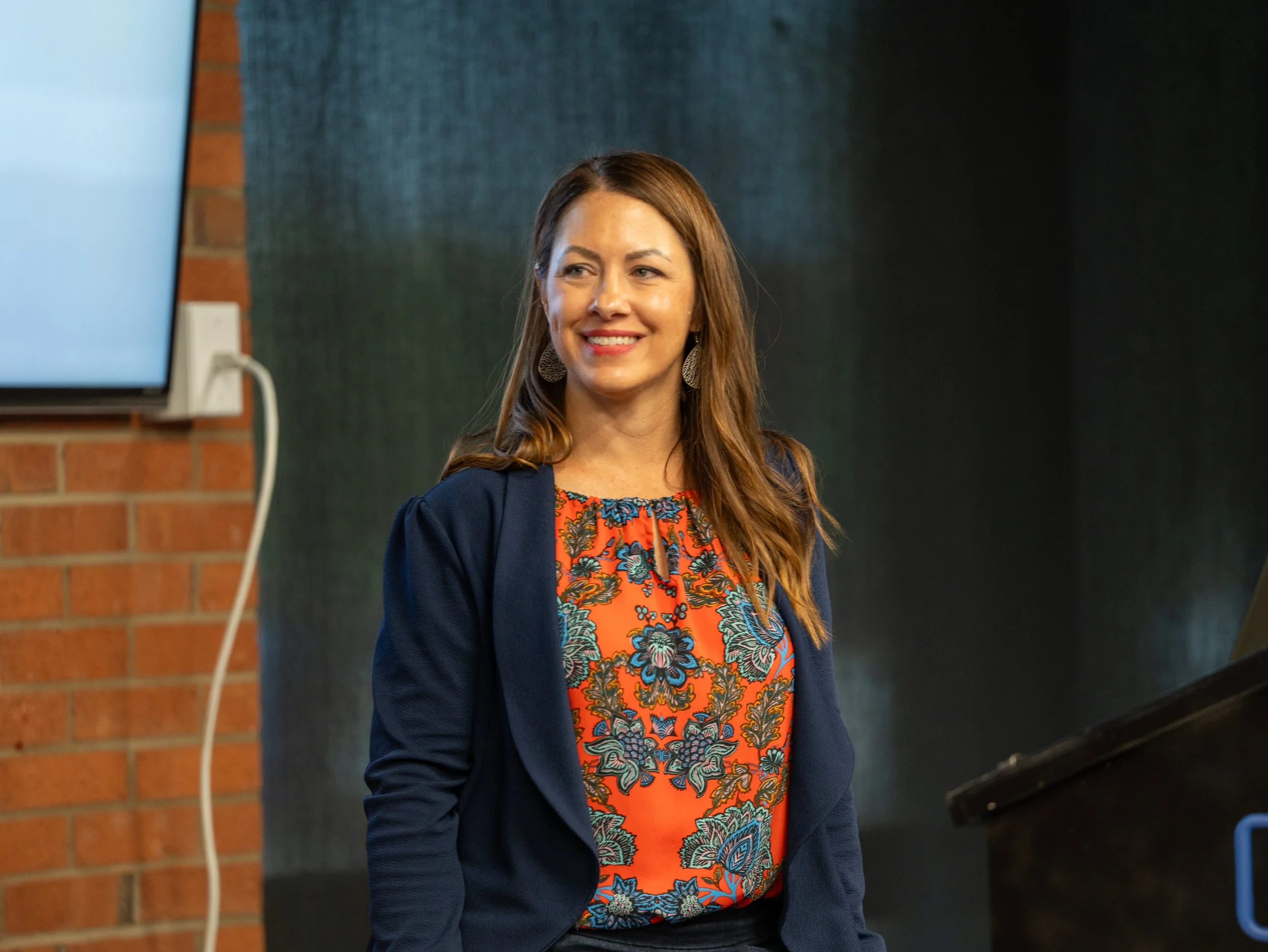 A woman with long brown hair, smiling, wearing earrings, a patterned orange top, and a dark blue jacket, standing near a brick wall and a black surface, with a monitor and a power outlet visible in the background.