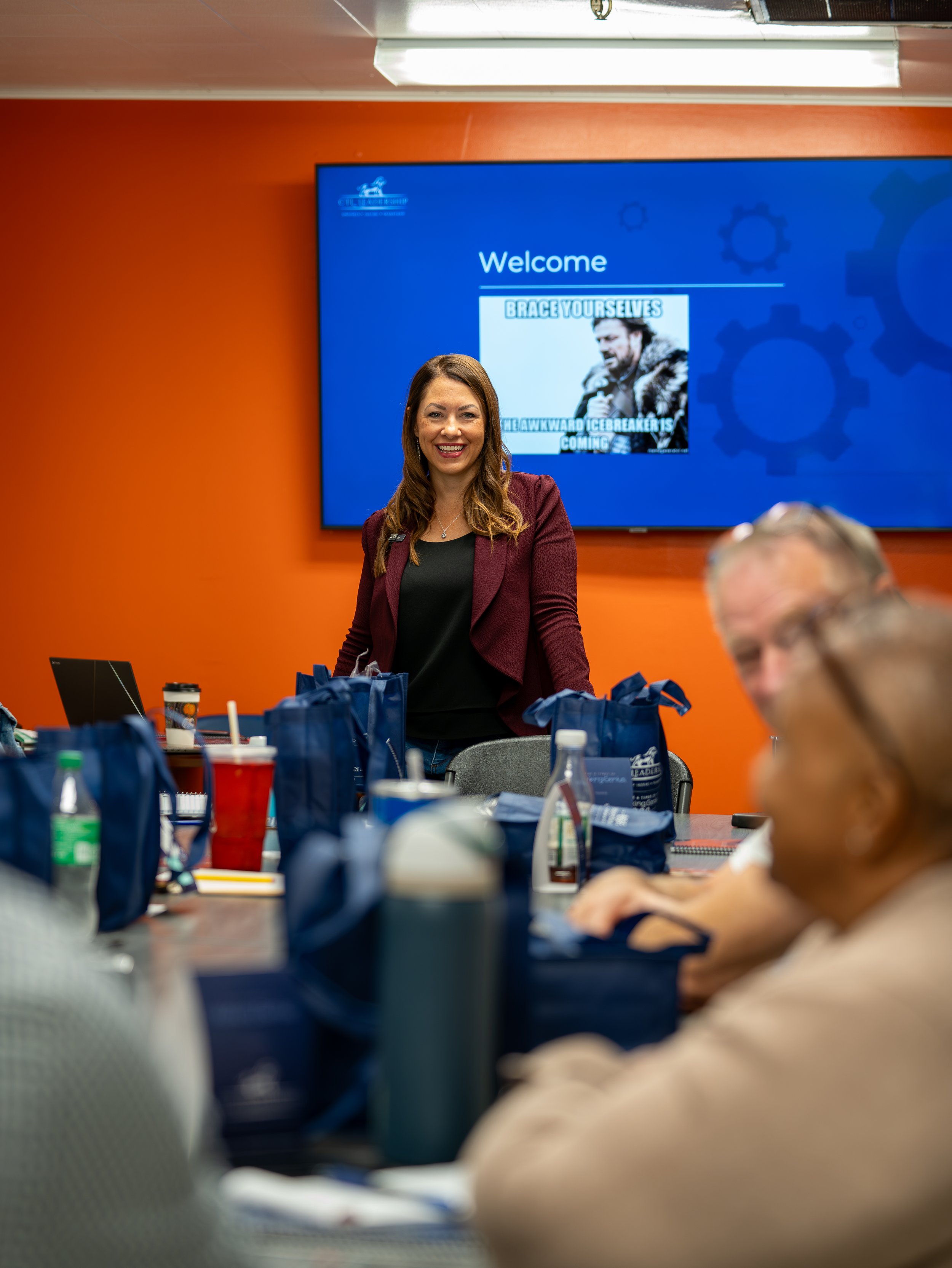 A woman in a maroon blazer smiling at a conference table with blue gift bags, drinks, and notebooks. A large screen behind her reads 'Welcome' with a meme showing a man in a fur coat with the text 'Brace yourselves, the awkward icebreaker is coming.'