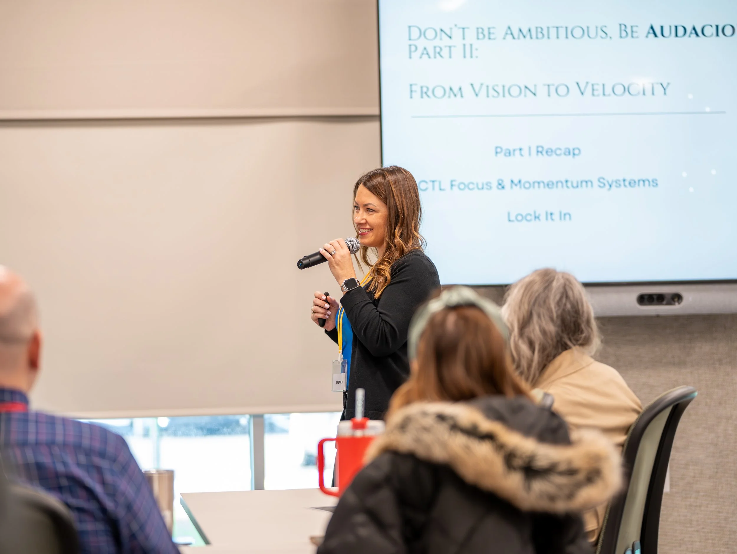 A woman with long brown hair holds a microphone and smiles as she presents in front of a screen displaying a PowerPoint slide. The slide has the title 'Don't be ambitious, be audacious' and mentions part II of a presentation, with the subtitle 'From Vision to Velocity.' Several audience members are visible, sitting at tables, some with notebooks and drinks.