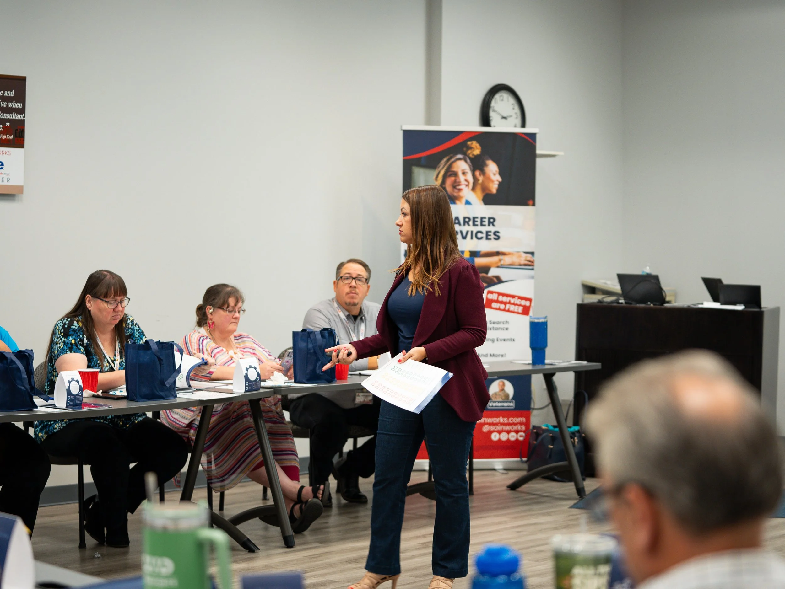 A woman with reddish-brown hair in a maroon blazer and blue jeans speaking to a group at a career and job fair. She is standing in front of a table with seated attendees, some wearing glasses, and a banner behind her displaying smiling women and the text "CAREER SERVICES."