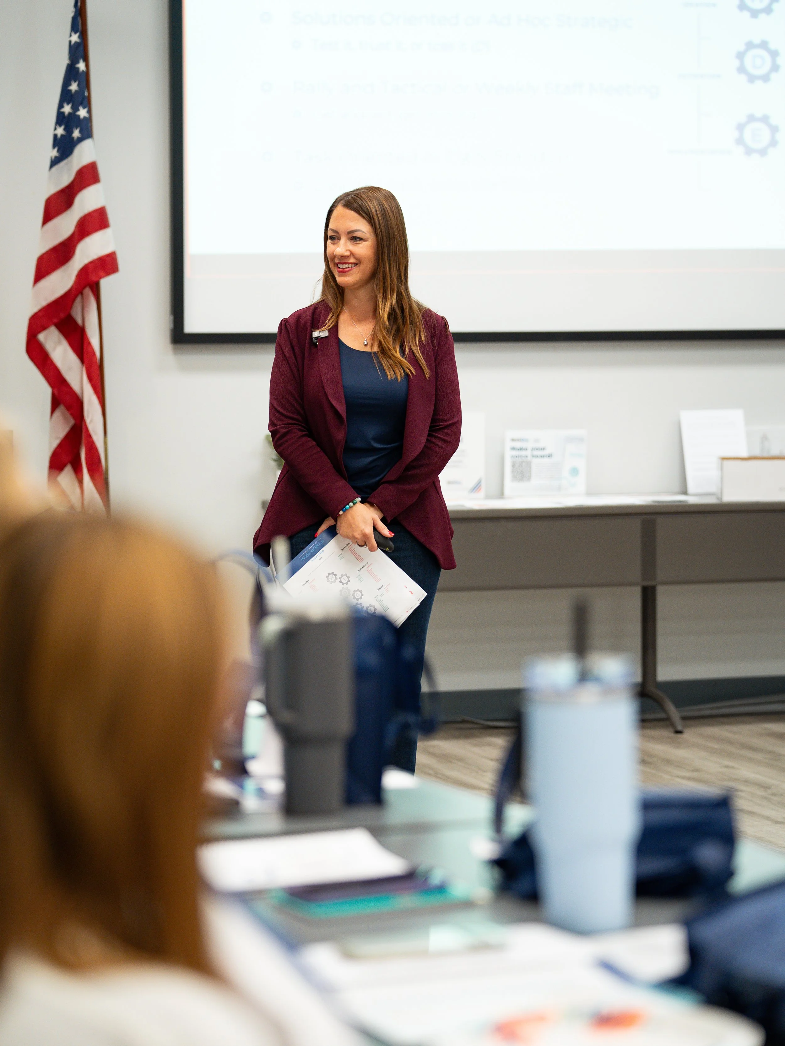 A woman with long brown hair smiling and holding papers in front of a projection screen in a conference room. An American flag is on the left side of the image, and there are display boards and tables with materials in the foreground.
