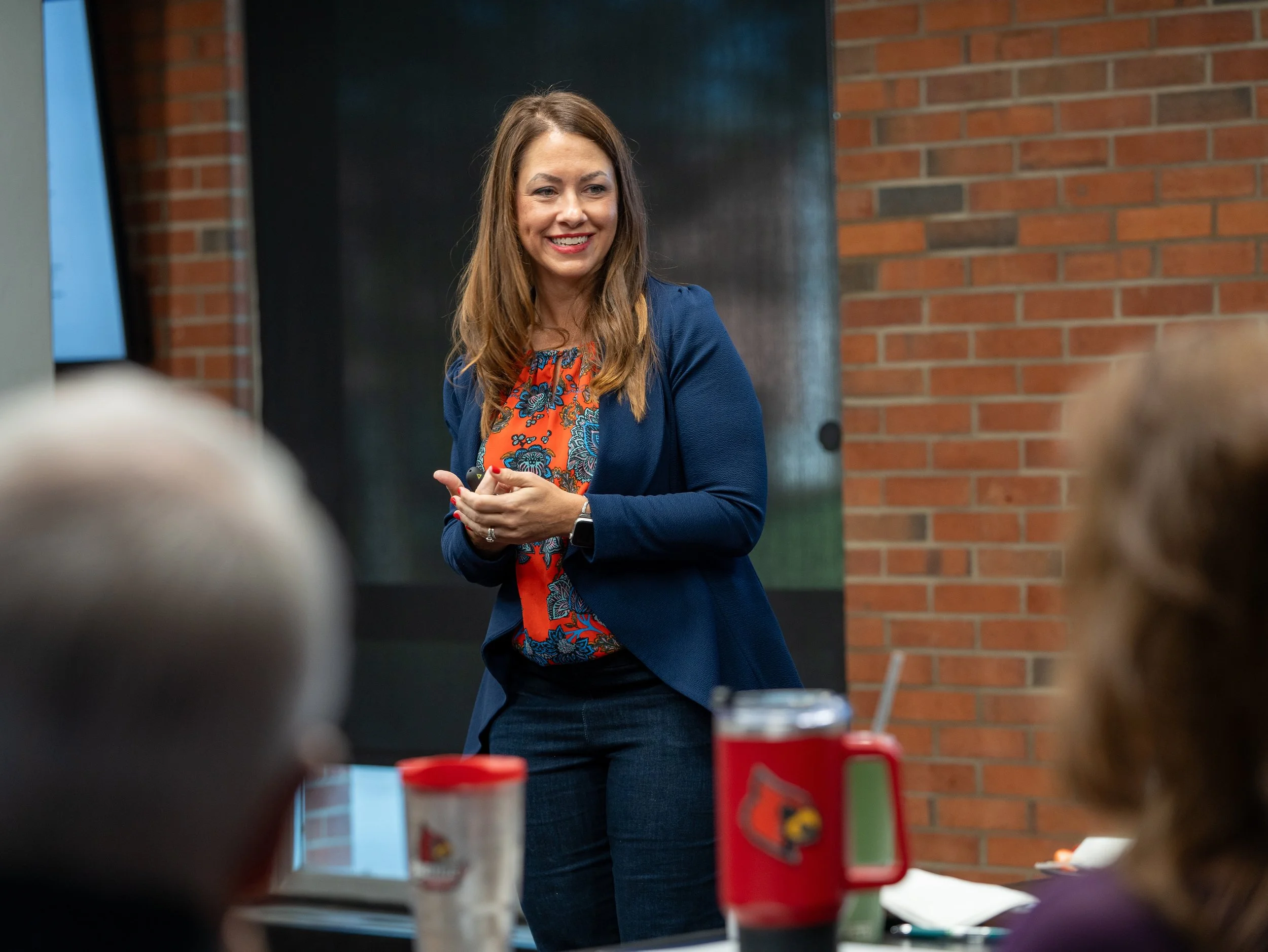A woman with long brown hair wearing a blue blazer and a colorful blouse stands smiling in front of a brick wall, speaking to an audience in a room with tables and food items.