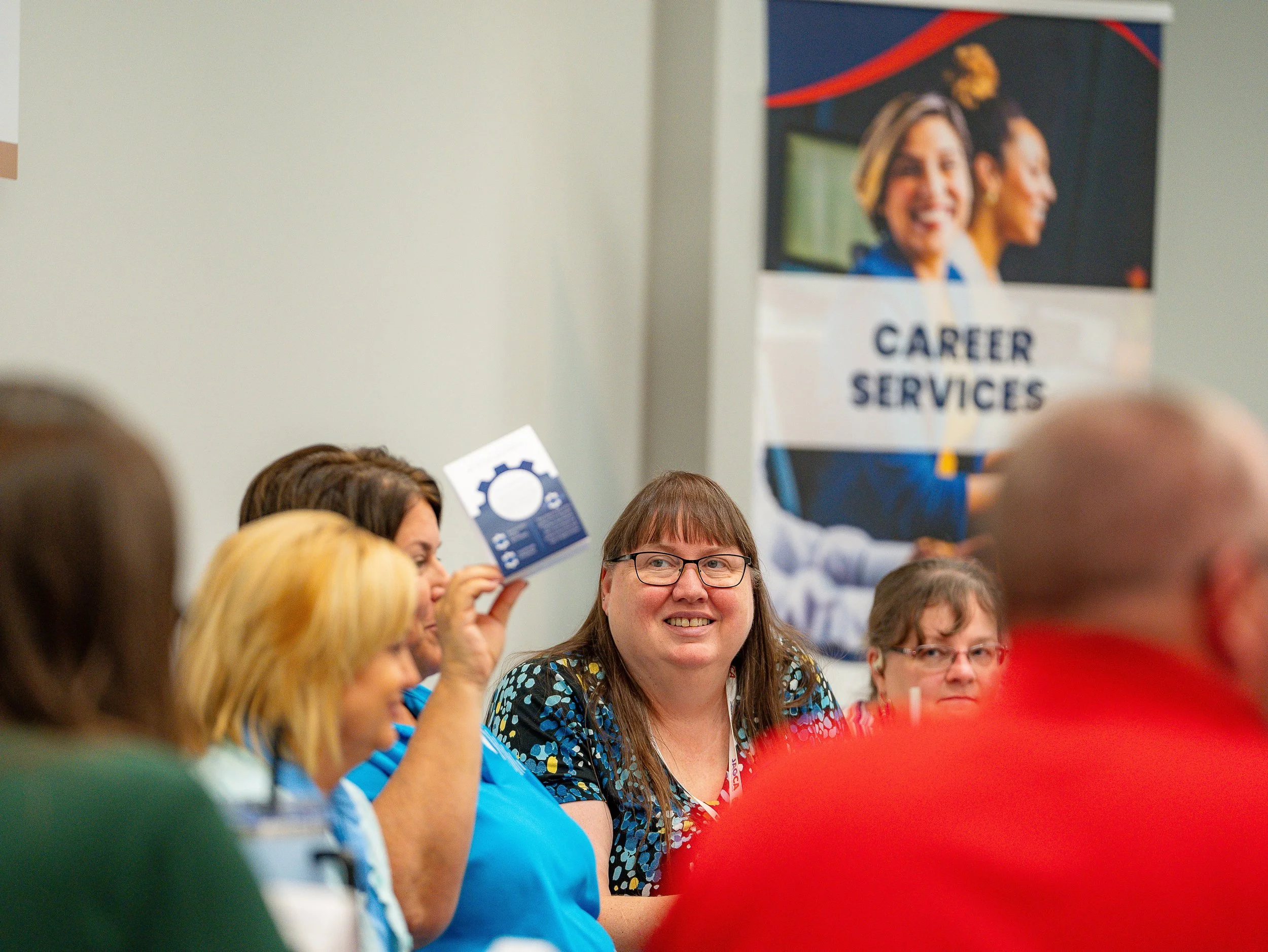 People seated at a table during a work or conference event, with a large poster in the background reading "Career Services" and showing smiling women.