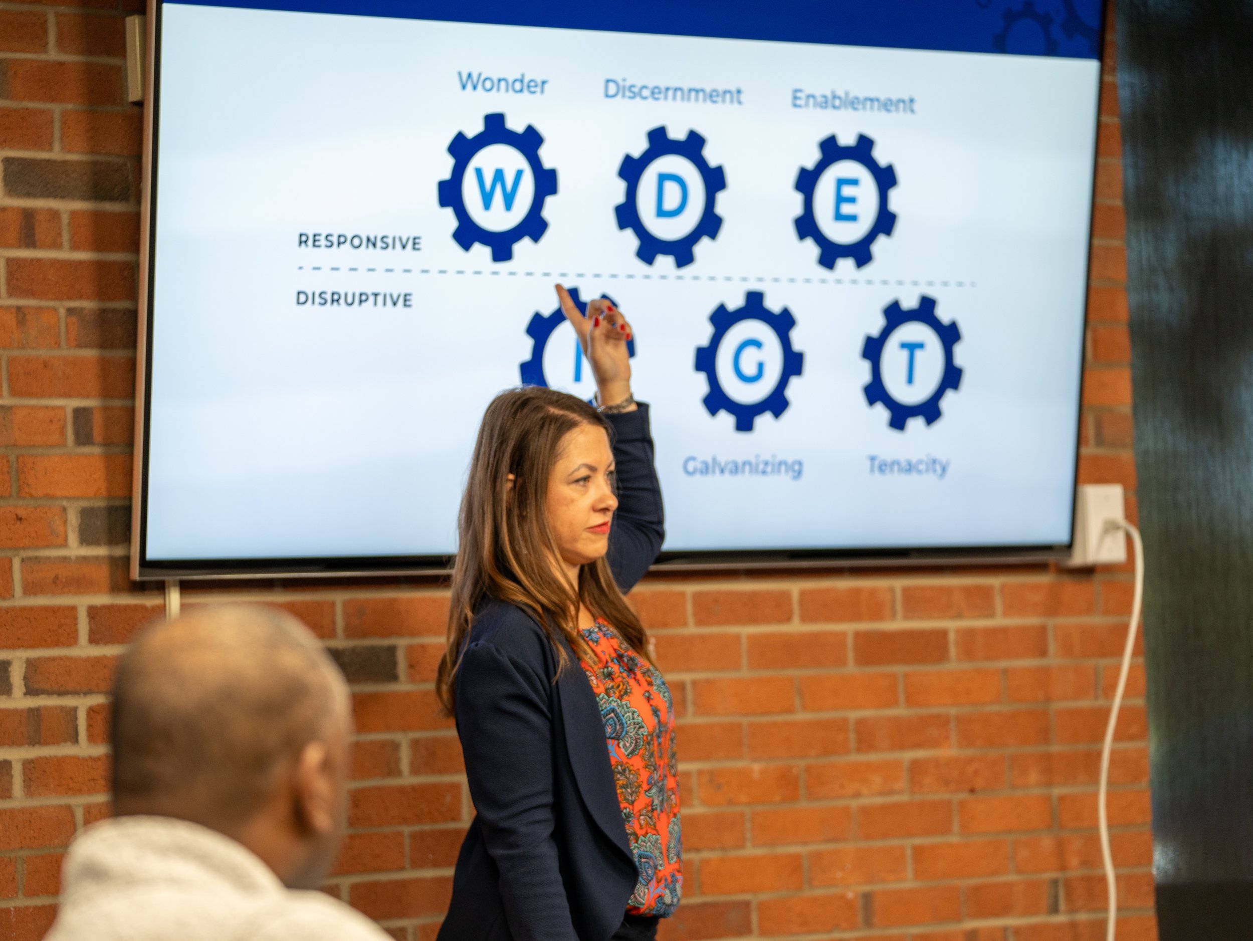 A woman standing in front of a presentation screen with gear icons labeled W, D, E, G, T. The screen has text related to different phases: Wonder, Discerning, Enablement, with additional words like Responsive, Disruptive, Galvanizing, Tenacity. The woman is wearing a dark blazer and colorful top, raising her right hand. The background features a brick wall.