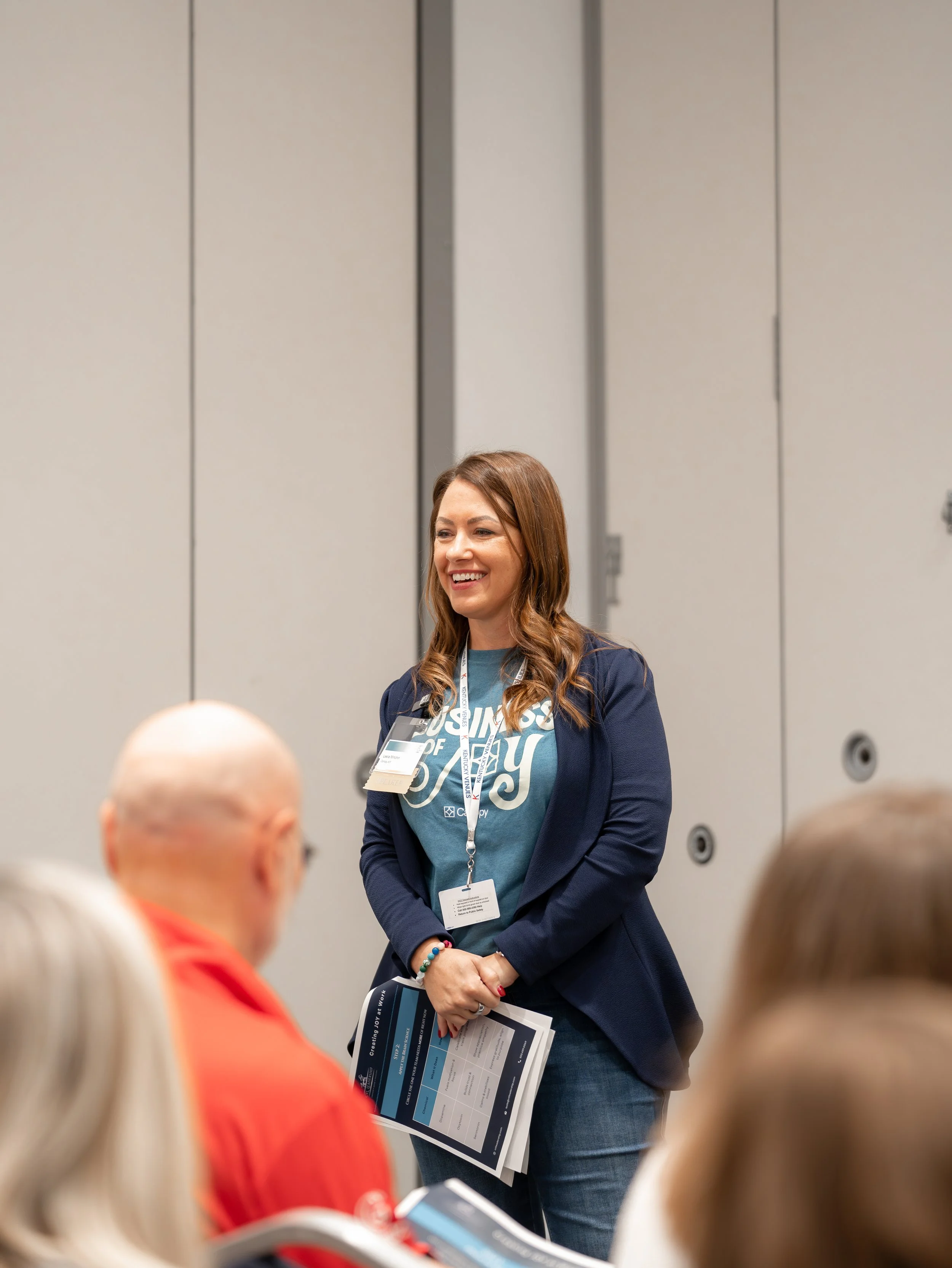 A woman with brown hair wearing a blue shirt and blazer smiling and talking to a group of people at a conference or seminar.