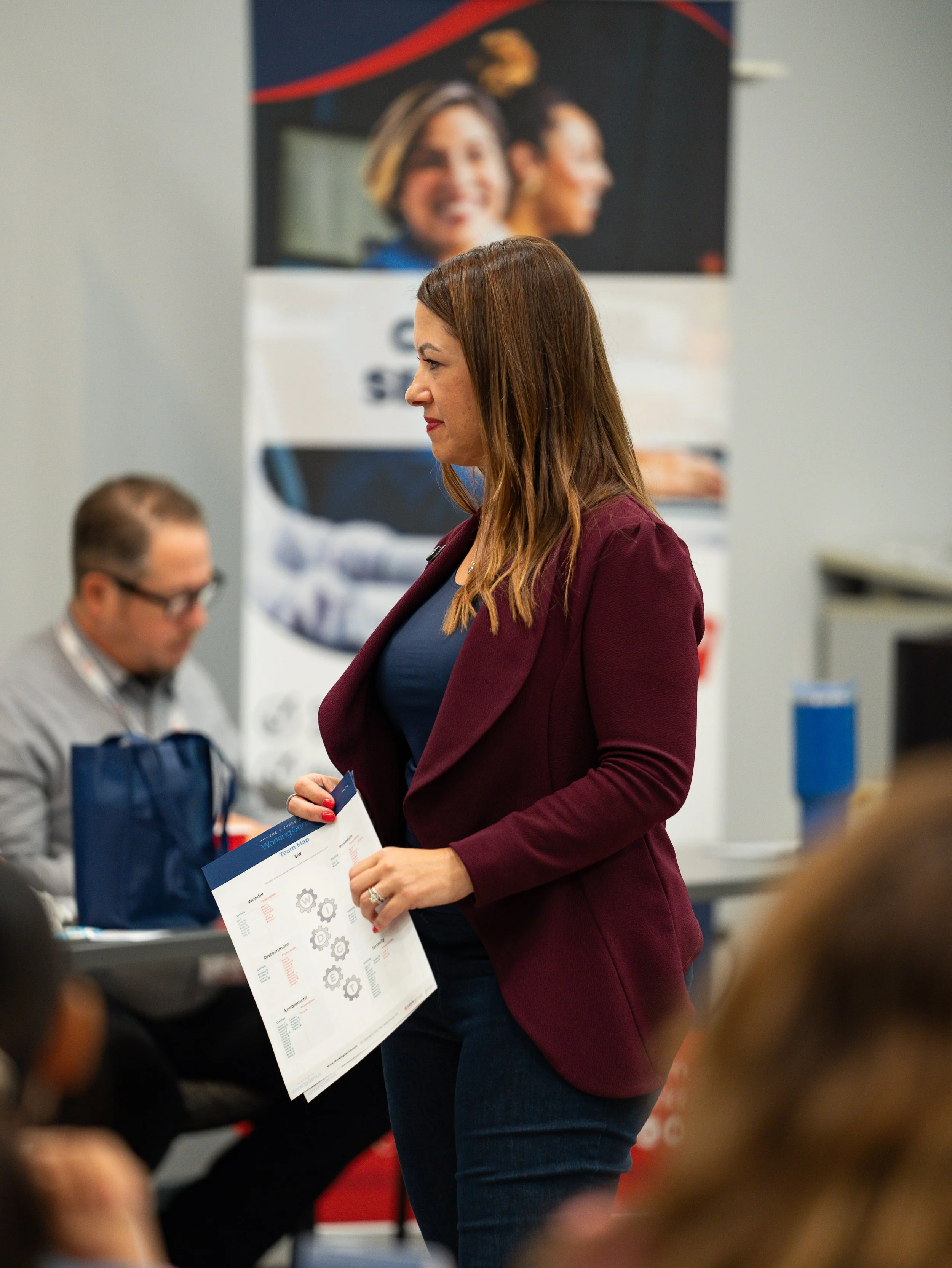 A woman with brown hair in a maroon blazer is holding a sheet with gears and charts, and is engaging with others at an indoor event. There is a blurred male participant with glasses and a lanyard in the background, and a poster with two smiling women behind her.