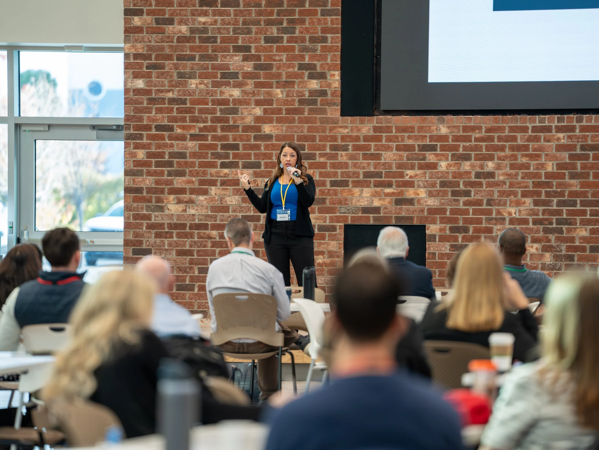 A woman in professional attire giving a presentation to an audience in a room with natural light, exposed brick walls, and a large screen.