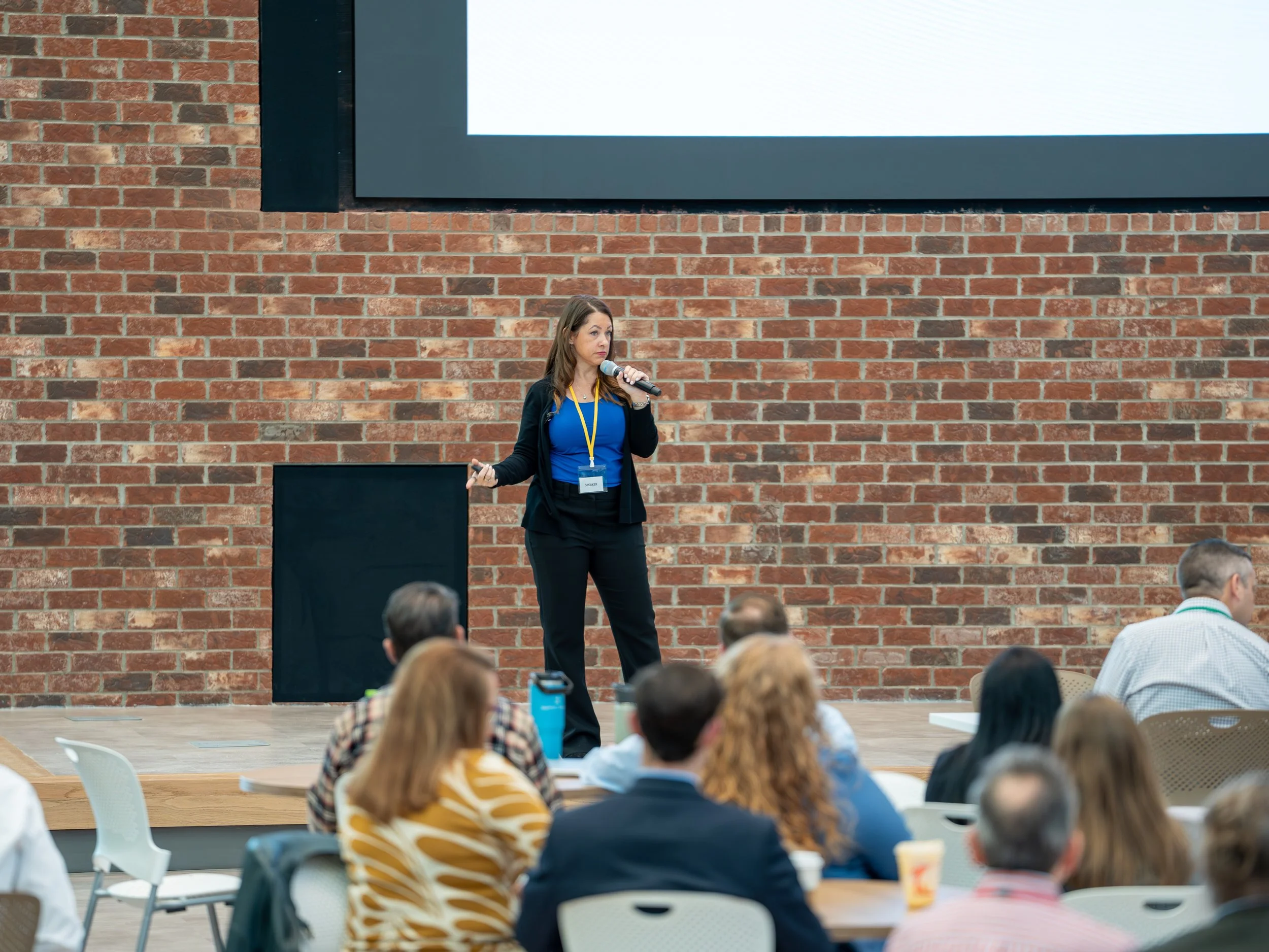A woman in business attire standing in front of a brick wall, speaking into a microphone and giving a presentation to an audience.
