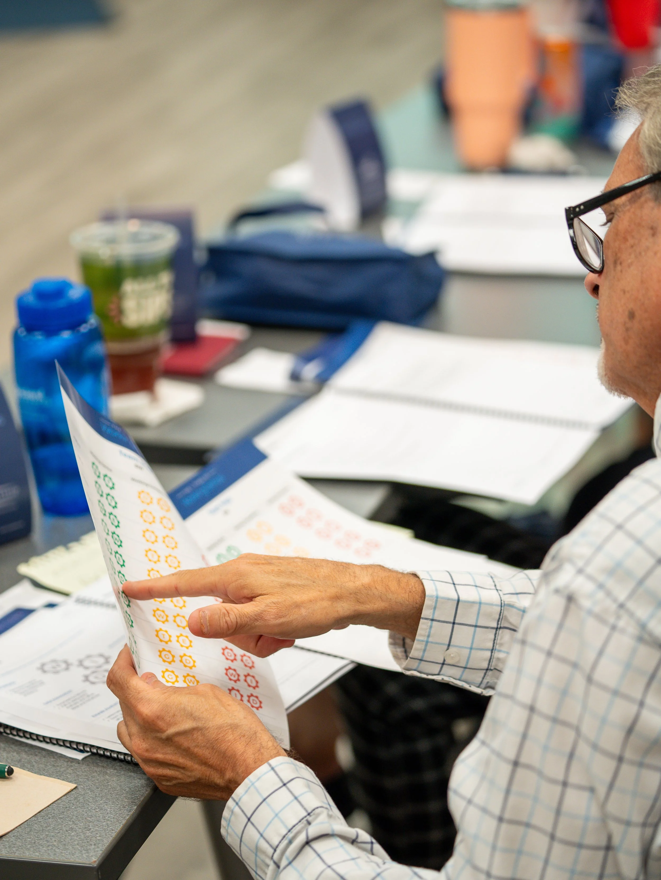Close-up of an older man with gray hair and glasses wearing a checked shirt reading a printed sheet with colorful icons, sitting at a desk with papers, folders, and a blue water bottle.