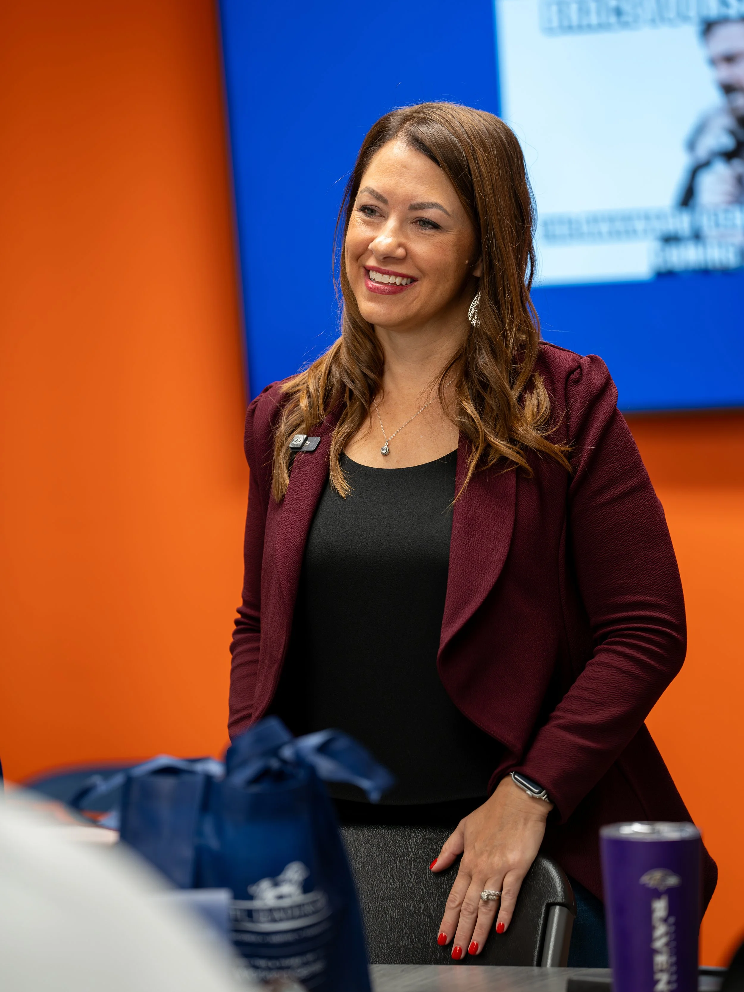 A woman with long brown hair, wearing a burgundy blazer and black top, smiling and sitting at a table in a conference room with orange and blue walls and a screen in the background.