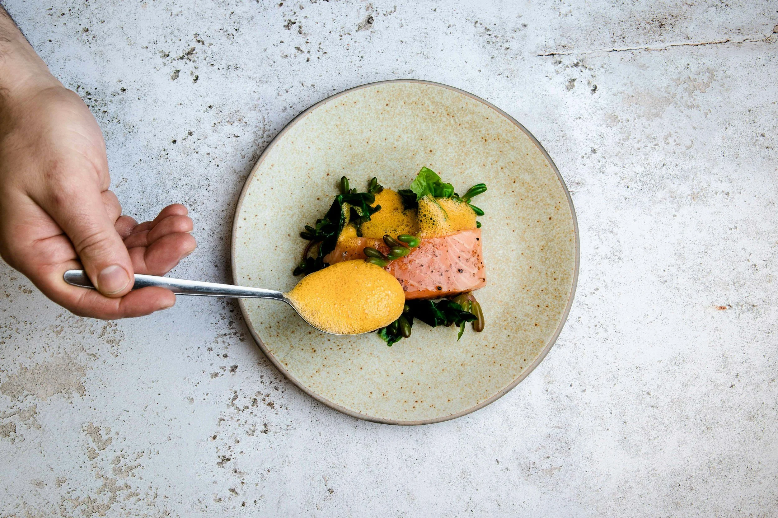 A plate of cooked salmon with green vegetables, garnished with foam sauce, on a speckled off-white surface.
