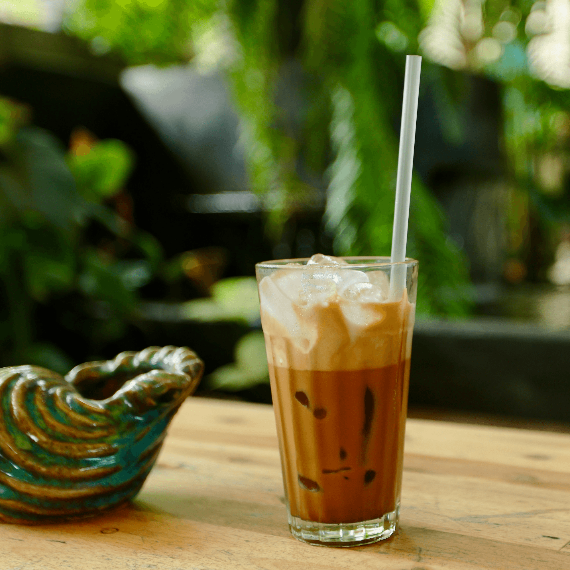 Close-up of a tall glass filled with iced coffee topped with whipped cream, with a white straw, on a wooden table with a green leafy background.