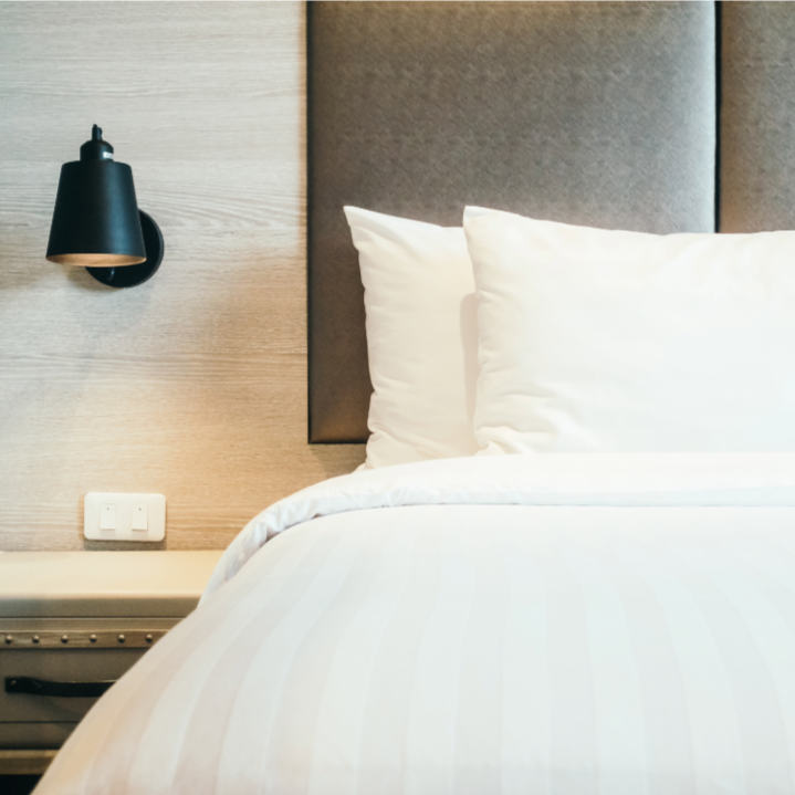 Close-up of a neatly made bed with white bedding and pillows in a modern hotel room, featuring a black wall-mounted lamp and a beige wall with a power outlet and bed headboard.