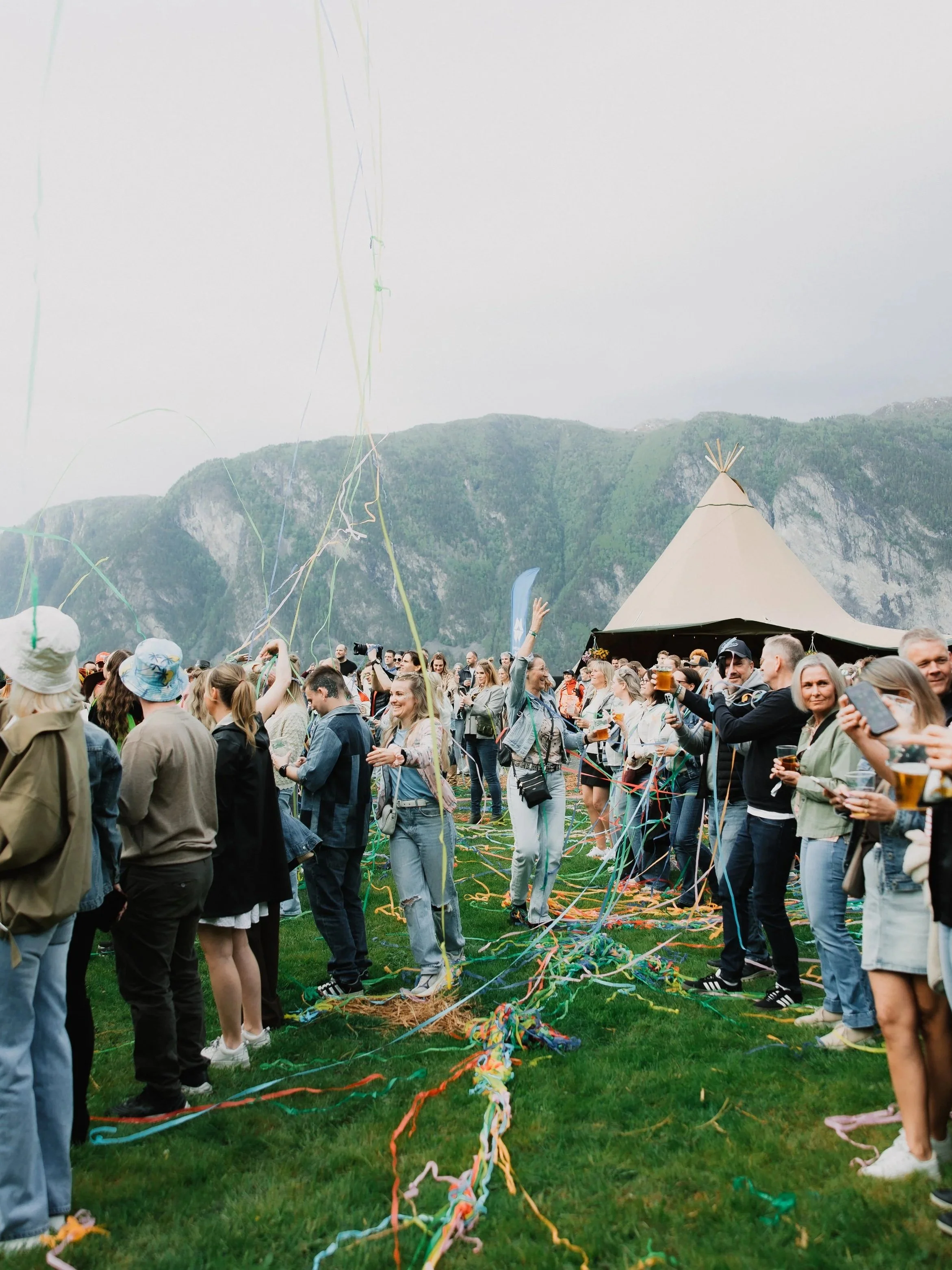 En stor gruppe mennesker samles utendørs på gresset under en festival eller et arrangement, med en tepee og fjell i bakgrunnen. Det er mange folk som smiler, tar bilder og har drikke i hånden, mens fargerike bånd og serpentiner flyr i luften.