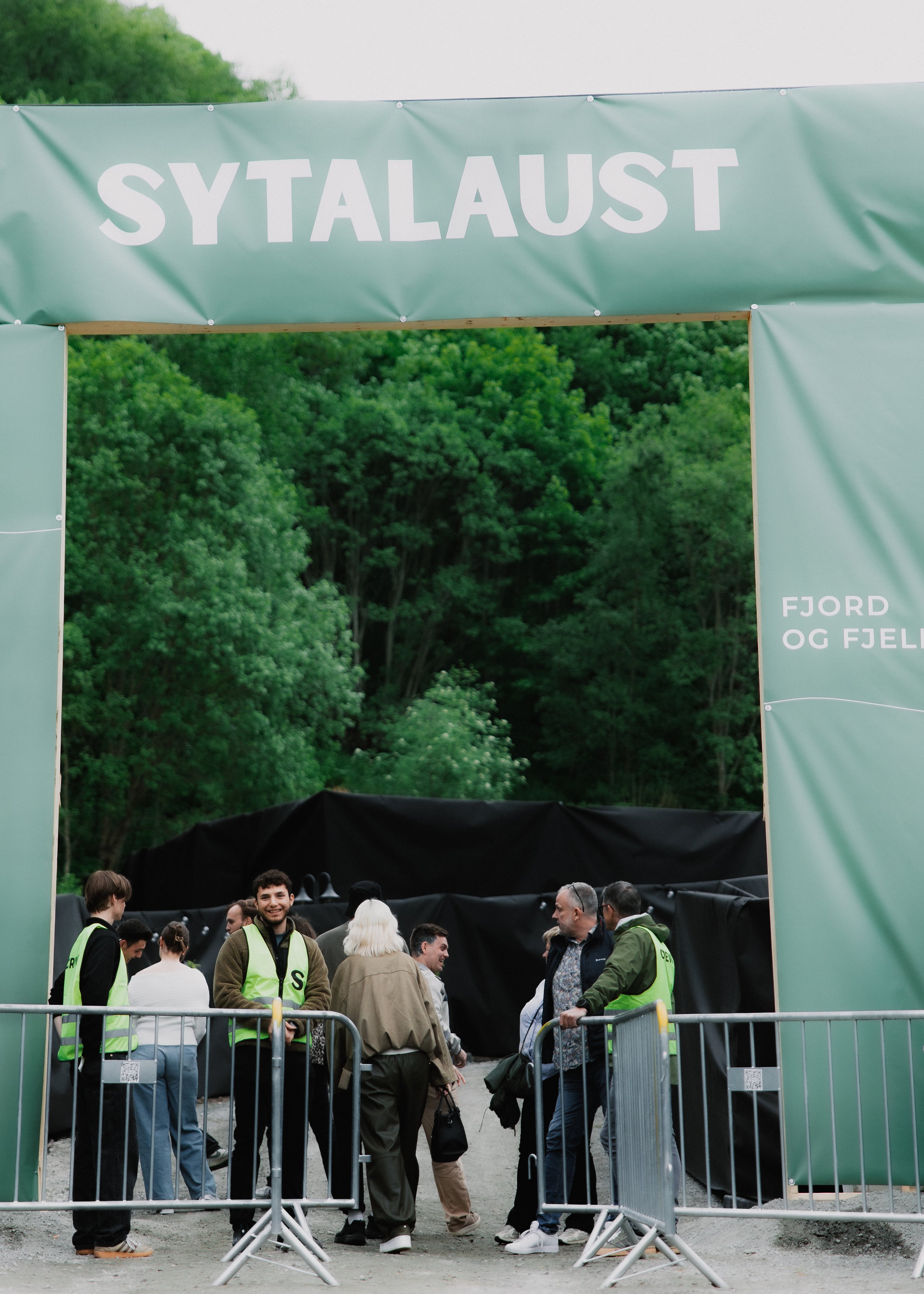 Mennesker står bak sperrebokker foran en inngangsport merket med skiltet 'Syttalaust' og bak en landlig skogkledd bakgrunn.