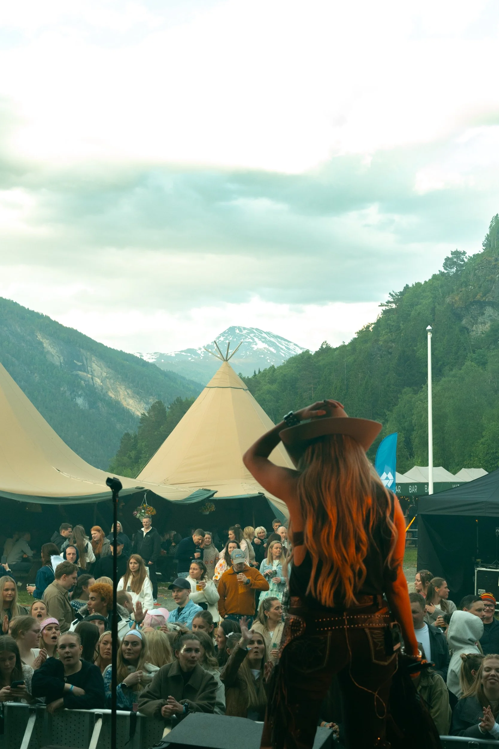 En person med langt rødt hår spiller på scene foran et stort folk som ser ut til å delta på et utendørs festival. Bakgrunnen viser fjell og grønne skoger, mange telt og en gruppe mennesker samlet foran scenen.